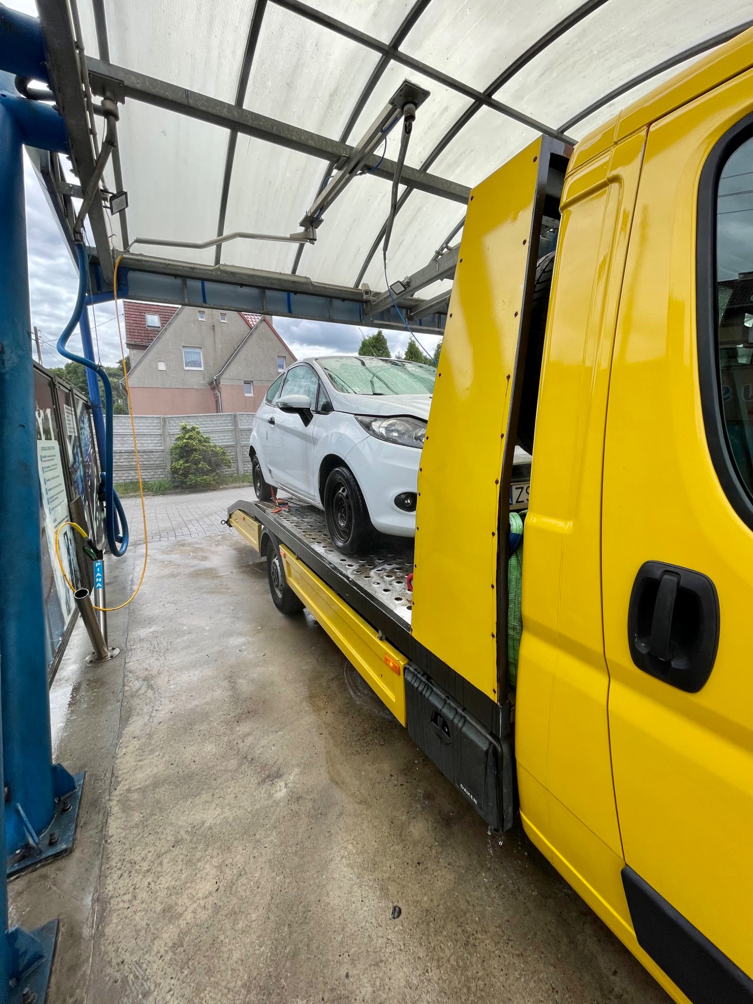 Żółta laweta z białym samochodem osobowym marki Ford na platformie, sfotografowana w myjni samochodowej z perspektywy lekko z dołu.