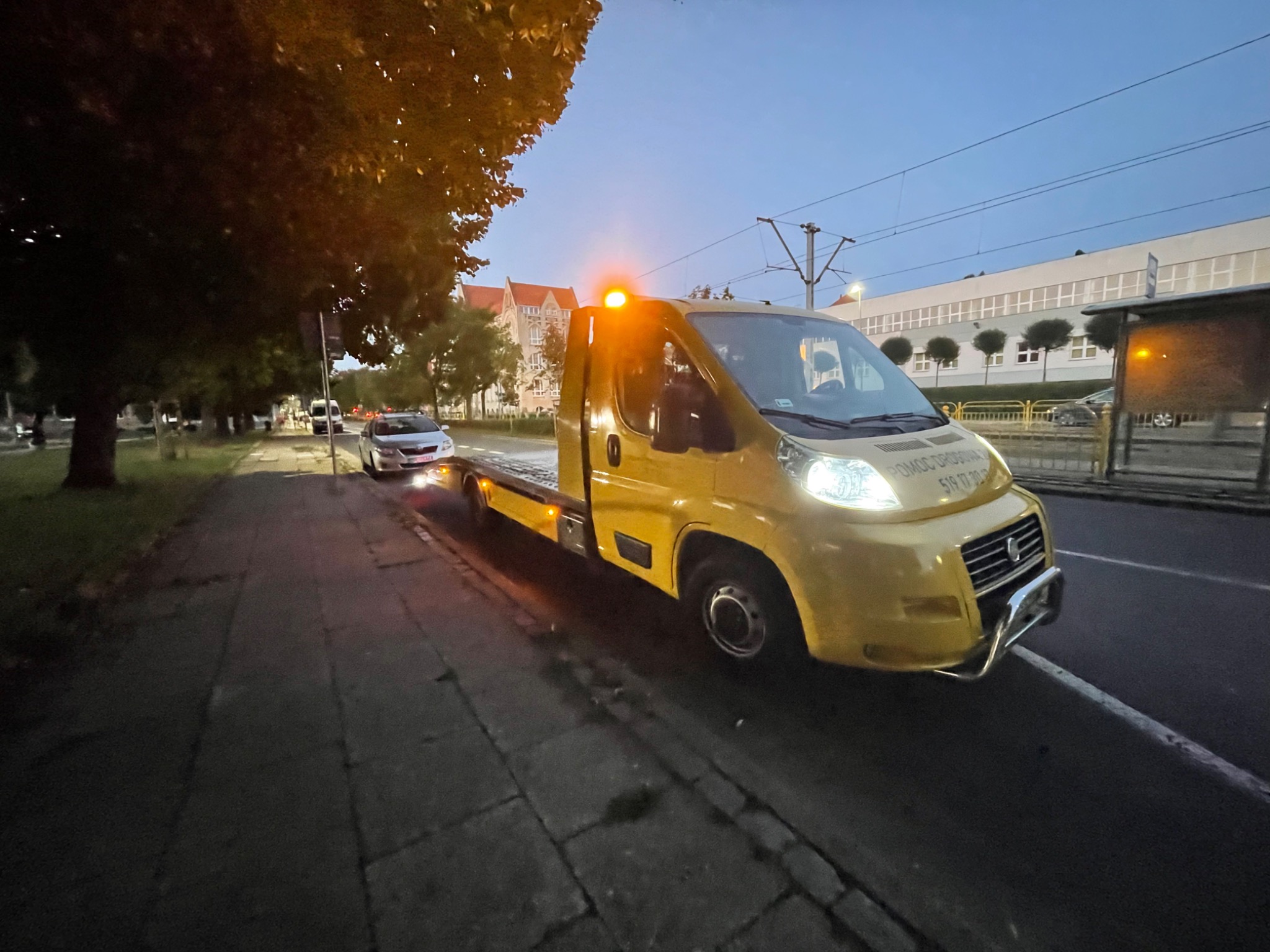 Żółta laweta z włączonym pomarańczowym światłem ostrzegawczym i reflektorami, zaparkowana na ulicy obok chodnika, w tle budynki i drzewa o zmierzchu.