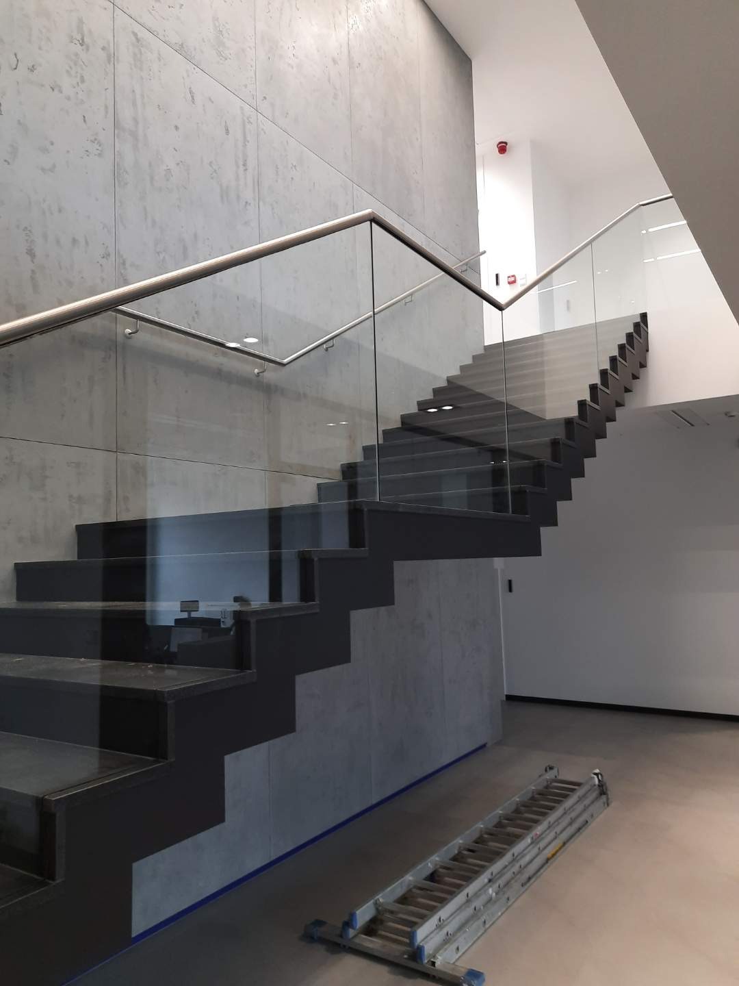Nowoczesne schody z czarnymi stopniami i szklaną balustradą z nierdzewnymi poręczami, widoczna rozłożona drabina na podłodze, szary beton architektoniczny na ścianie.
