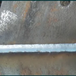 Szlif spoiny na złączu dwóch metalowych elementów o surowej, chropowatej powierzchni, widoczne ślady korozji i obróbki.