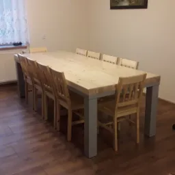 Duży, surowy drewniany stół z szarymi metalowymi nogami i ośmioma drewnianymi krzesłami w jasnym wnętrzu z brązową podłogą.