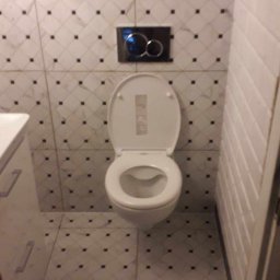 KLAR ARTUR MACIĄGA - Toaleta wisząca z otwartą deską, czarny przycisk spłuczki na tle białych płytek imitujących cegłę i płytek podłogowych z czarnymi kwadratami.