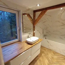 Jasna łazienka z drewnianymi akcentami: blat z drewna przy oknie, drewniana belka stropowa, płytki imitujące marmur, podłoga ułożona w jodełkę, biała umywalka nablatowa i bateria.