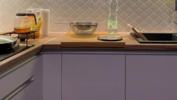 Fragment kuchni z jasnofioletowymi szafkami, drewnianym blatem, metalową miską, butelką wody i czarnym zlewem z drewnianymi przyborami w środku, na ścianie mozaika w odcieniach szarości.