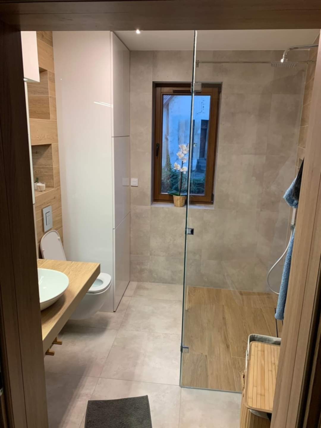 Nowoczesna łazienka z drewnianymi akcentami: blat umywalki z jasnego drewna, płytki imitujące drewno w strefie prysznica, szklana kabina i okno z widokiem na zieleń.