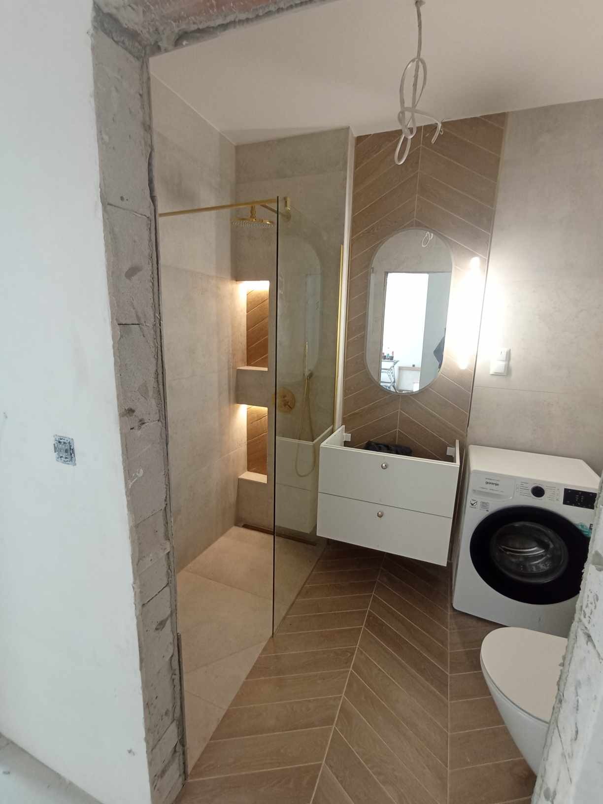 Nowoczesna łazienka w trakcie wykańczania, z kabiną prysznicową ze złotymi detalami, umywalką z szafką, pralką i toaletą, wykończona płytkami ułożonymi w jodełkę.