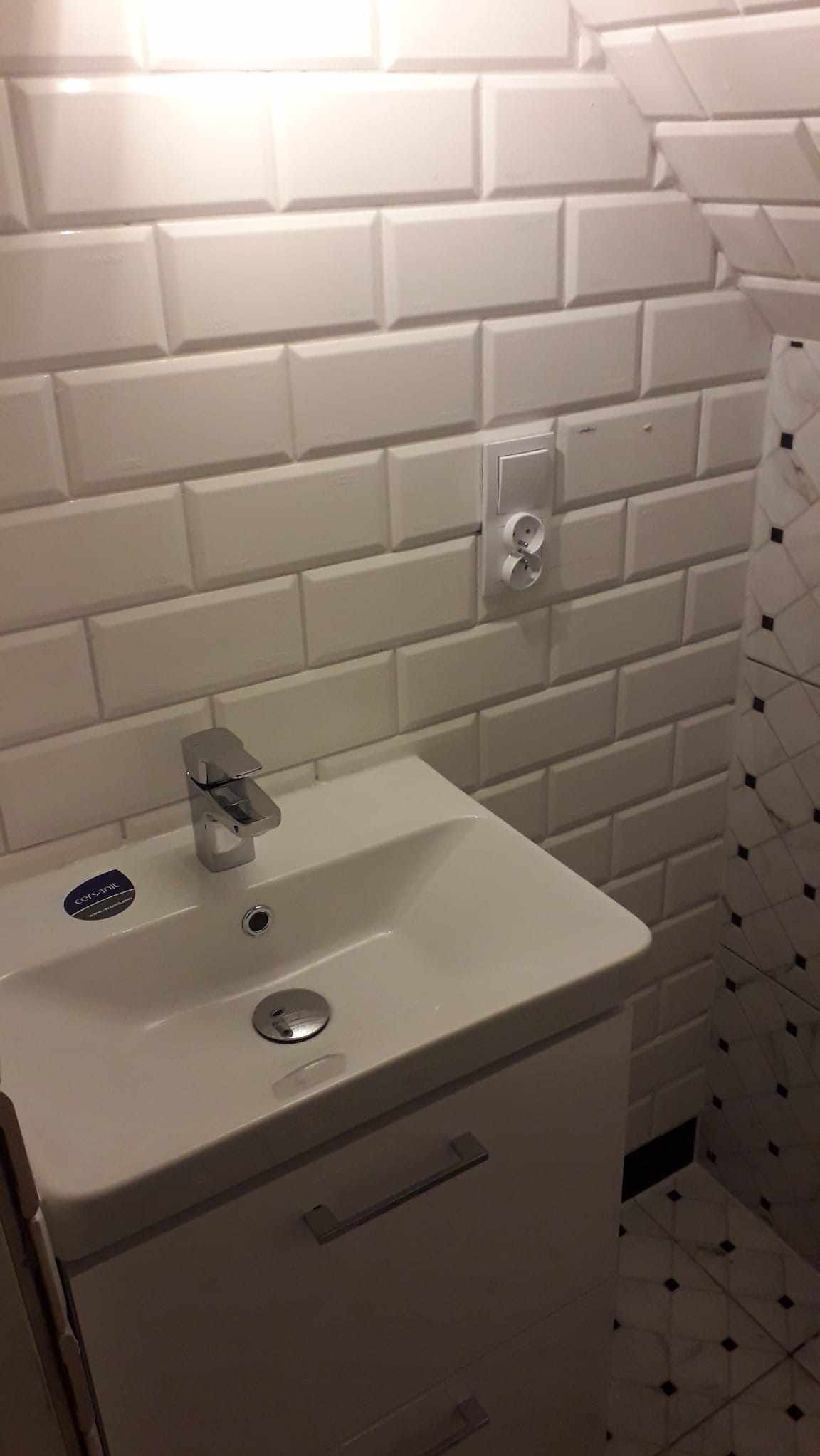 Biała umywalka z szafką i chromowaną baterią na tle ściany z białych, prostokątnych płytek, z widocznym gniazdkiem elektrycznym i fragmentem ściany wykończonej płytkami imitującymi marmur.