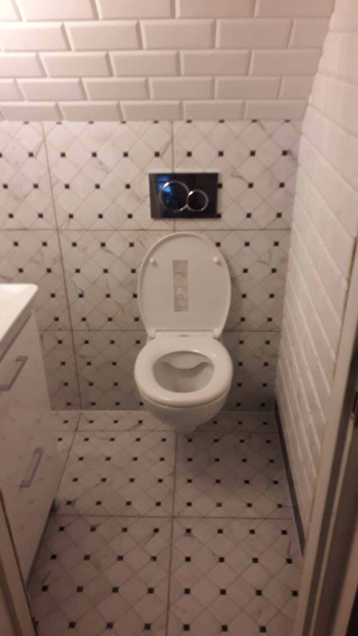 Toaleta wisząca z otwartą deską, czarny przycisk spłuczki na tle białych płytek imitujących cegłę i płytek podłogowych z czarnymi kwadratami.