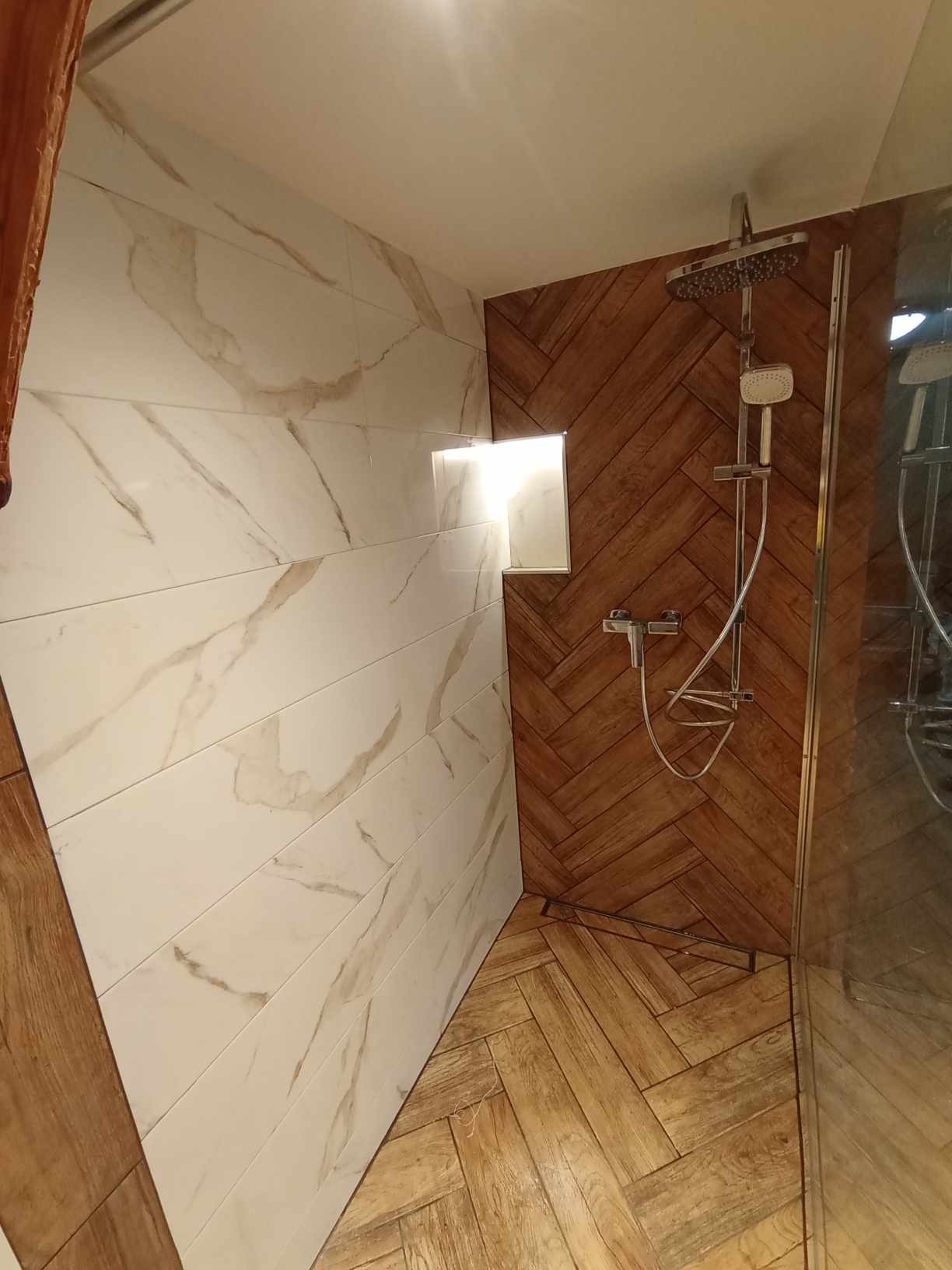 Wykończona łazienka z białymi płytkami imitującymi marmur na ścianie, drewnopodobnymi płytkami ułożonymi w jodełkę w strefie prysznica i na podłodze, oraz wnęką z oświetleniem.