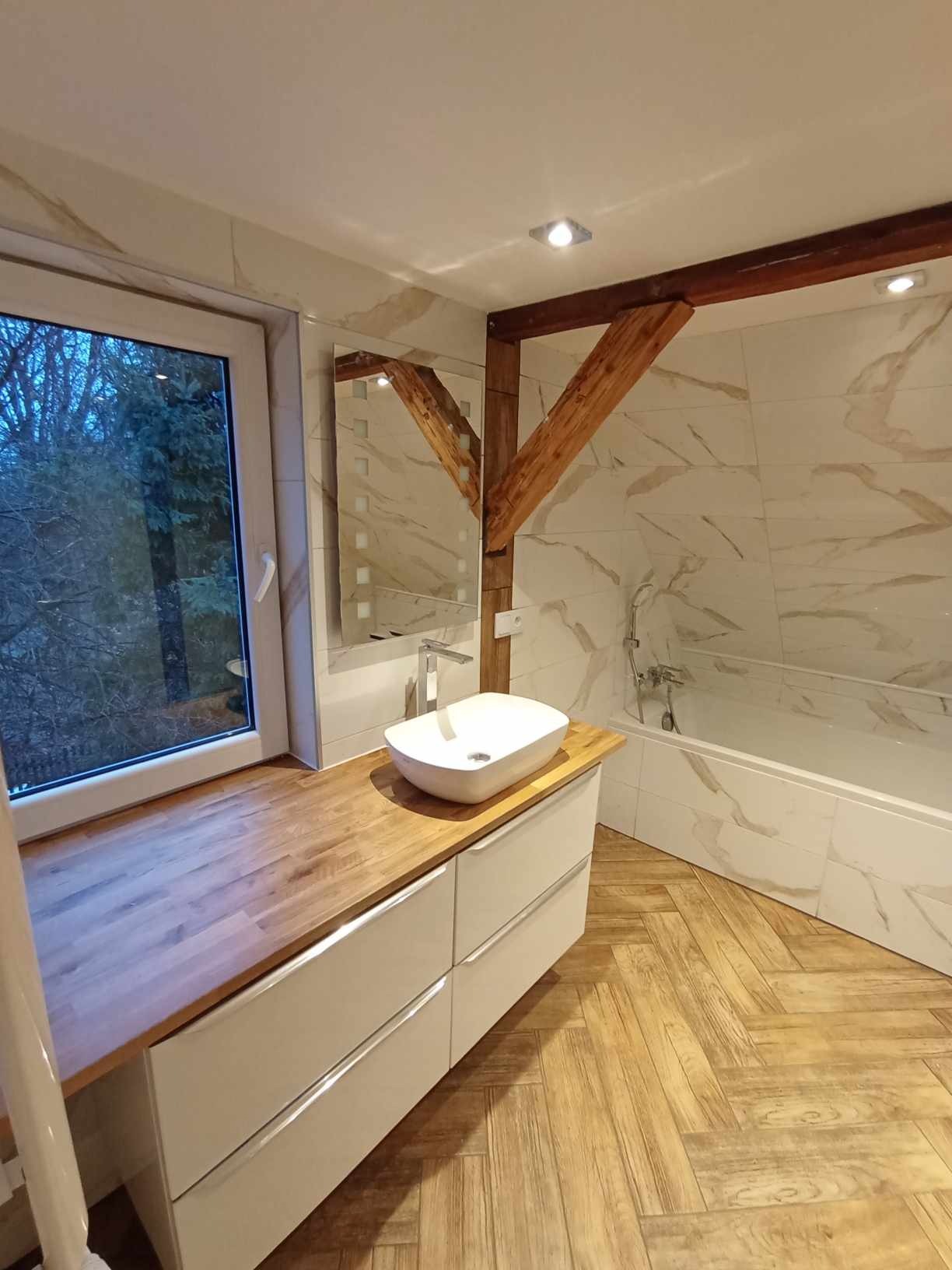 Jasna łazienka z drewnianymi akcentami: blat z drewna przy oknie, drewniana belka stropowa, płytki imitujące marmur, podłoga ułożona w jodełkę, biała umywalka nablatowa i bateria.