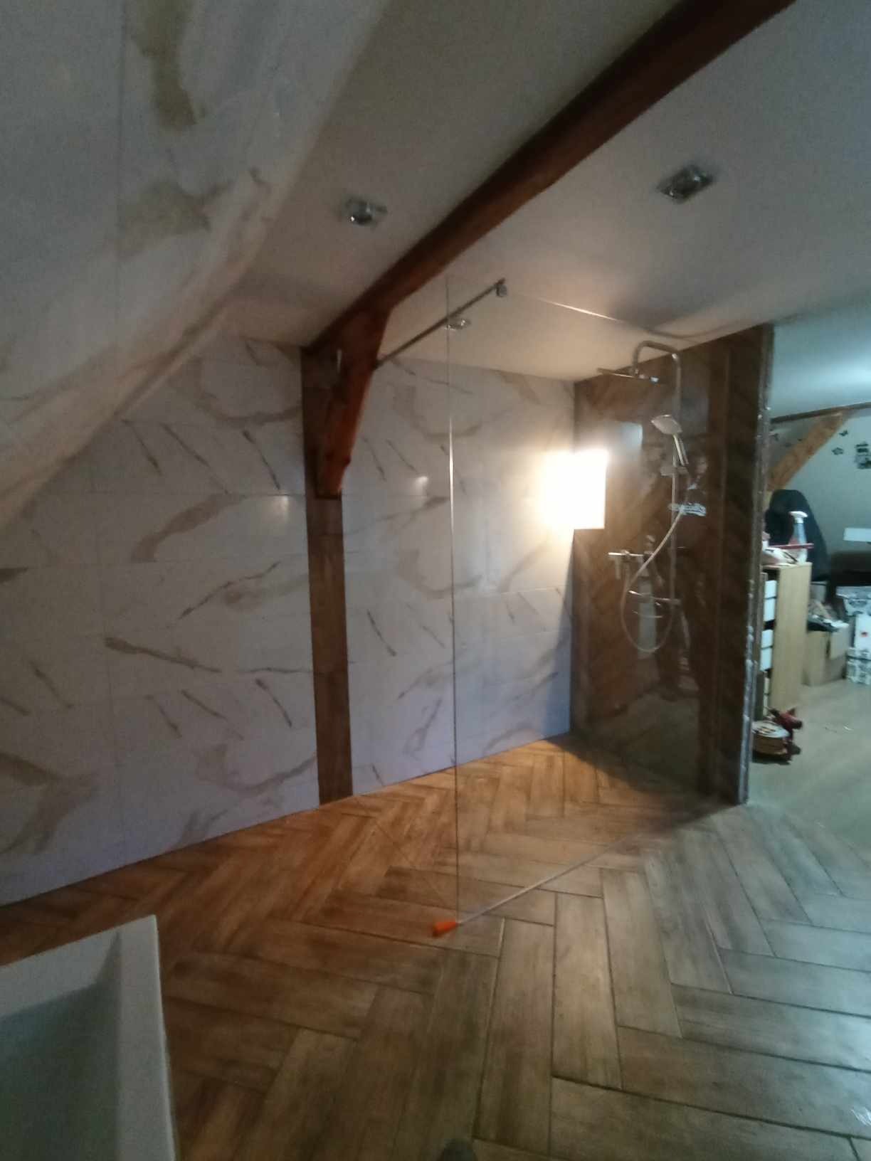 Nowoczesna łazienka na poddaszu z kabiną prysznicową typu walk-in, wykończona płytkami imitującymi marmur na ścianach i drewnianą jodełką na podłodze, z widoczną drewnianą belką stropową.
