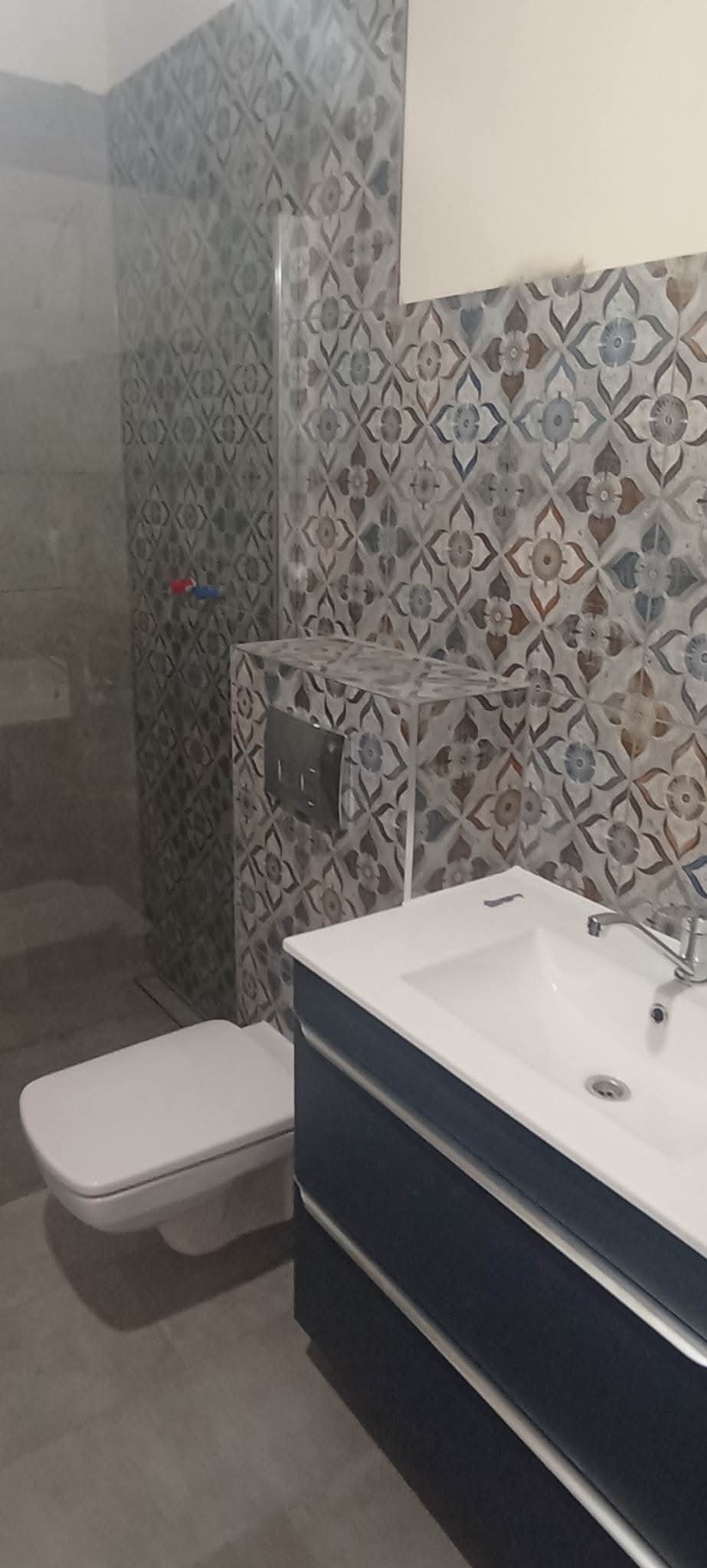 Wykończona łazienka z toaletą wiszącą, umywalką nablatową na szafce w kolorze granatowym oraz dekoracyjnymi płytkami ściennymi w odcieniach szarości, brązu i błękitu.