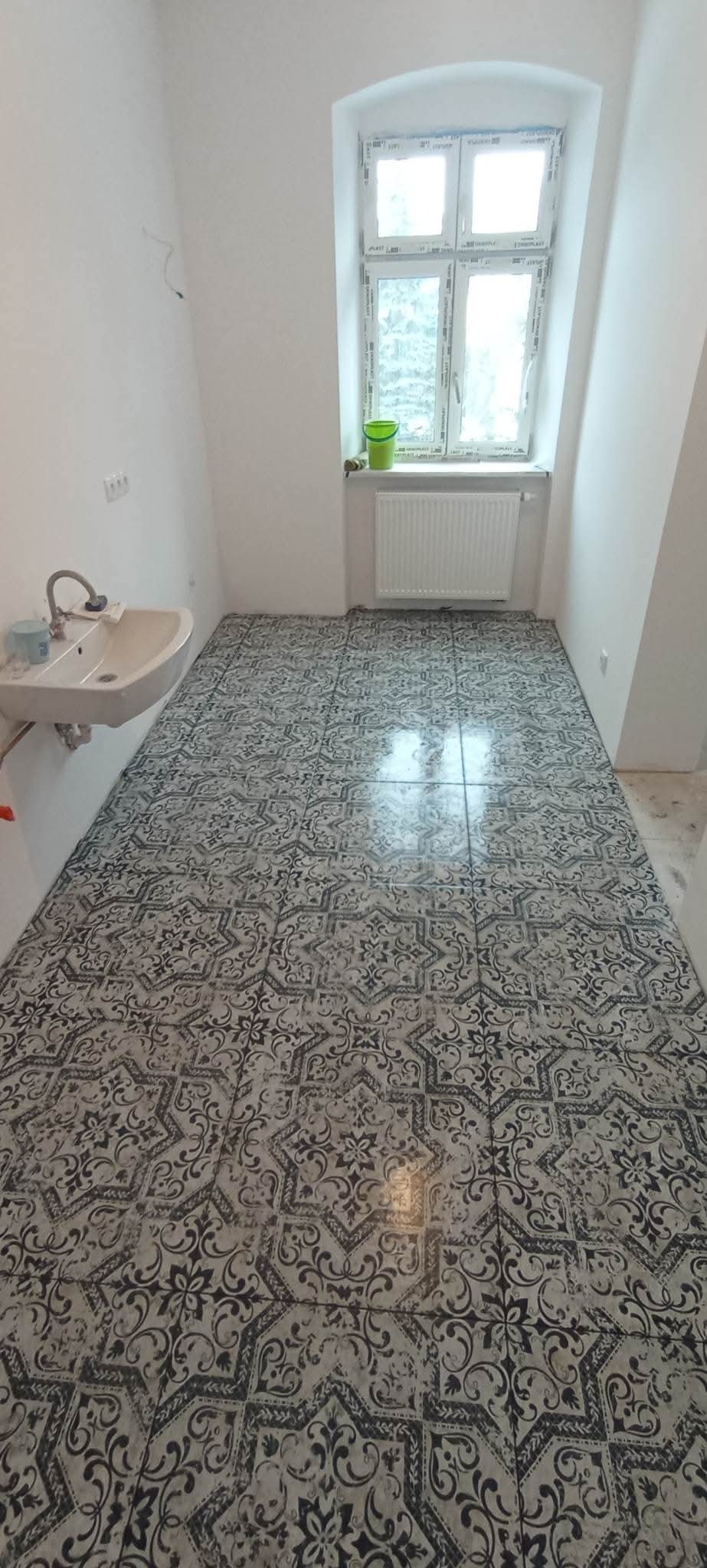 Ułożona podłoga z płytek w łazience z wzorem ornamentowym, umywalka i okno z grzejnikiem w tle.
