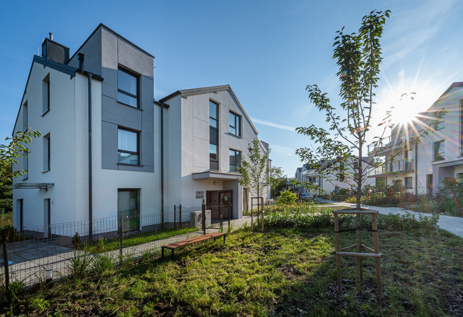 Nowoczesny budynek mieszkalny z elementami szarości, otoczony zielenią i młodymi drzewami. Słoneczny dzień, błękitne niebo. Widoczna ławka i ogrodzenie.