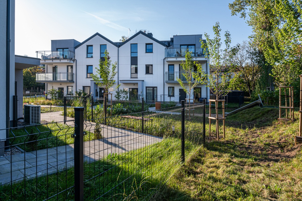 Nowoczesny budynek wielorodzinny z balkonami, otoczony zielenią i ogrodzeniem. Widoczne młode drzewka i trawnik. Słoneczny dzień.