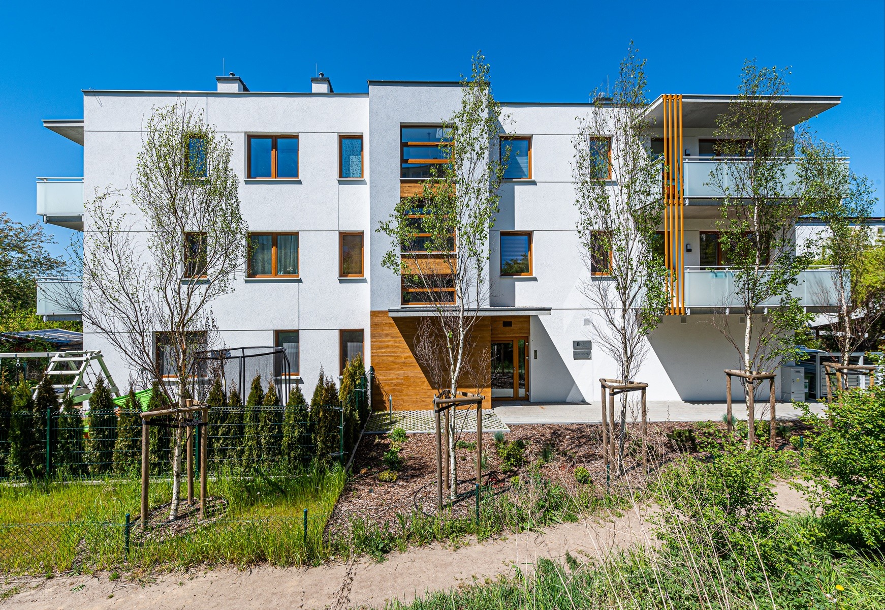 Nowoczesny, trzypiętrowy budynek mieszkalny z białymi ścianami, drewnianymi oknami i balkonami. Na pierwszym planie młode drzewa i zadbana zieleń.