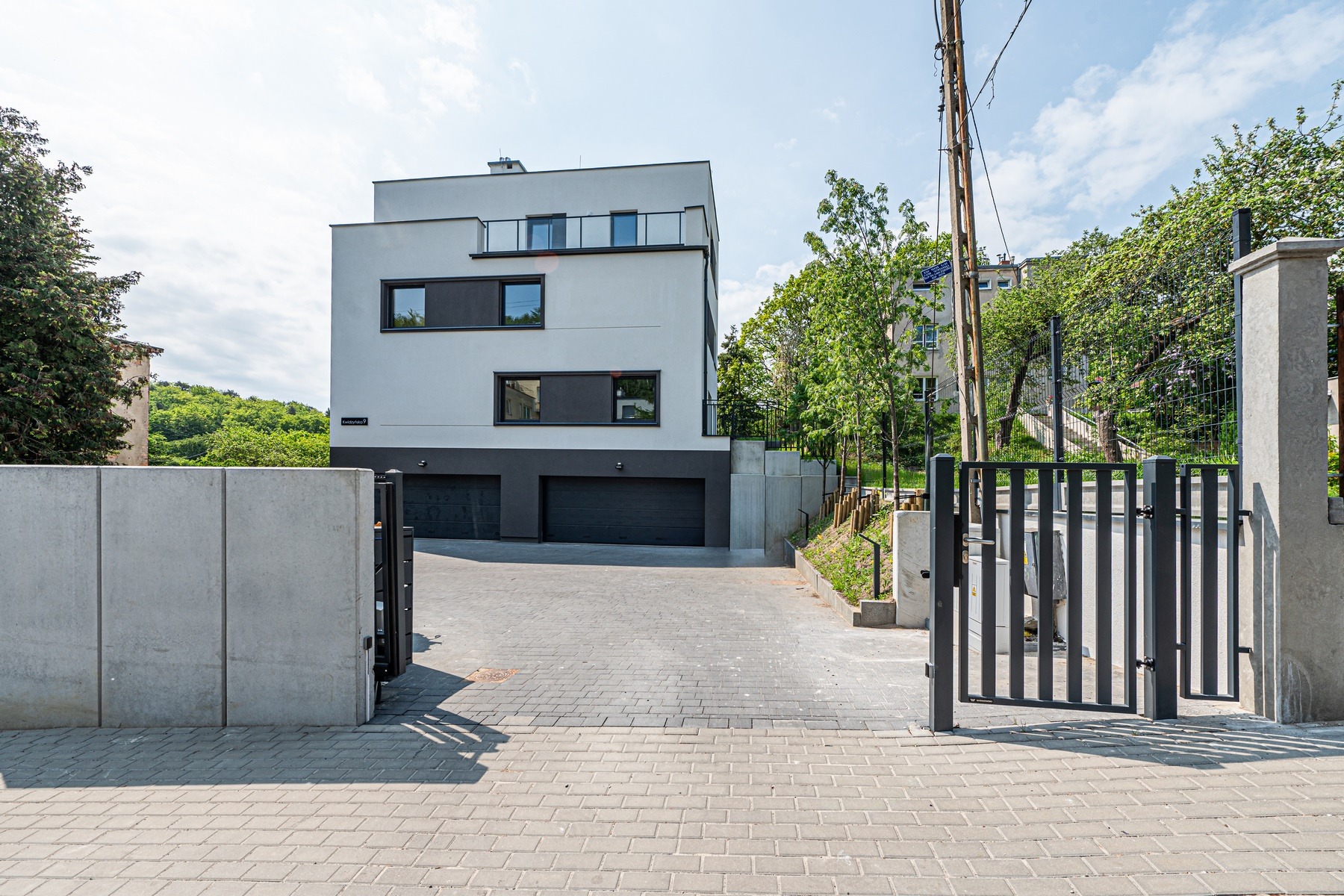 Nowoczesny dom z garażem w Gdyni, widok z zewnątrz przez otwartą bramę. Minimalistyczna architektura, szara kostka brukowa i betonowe ogrodzenie.