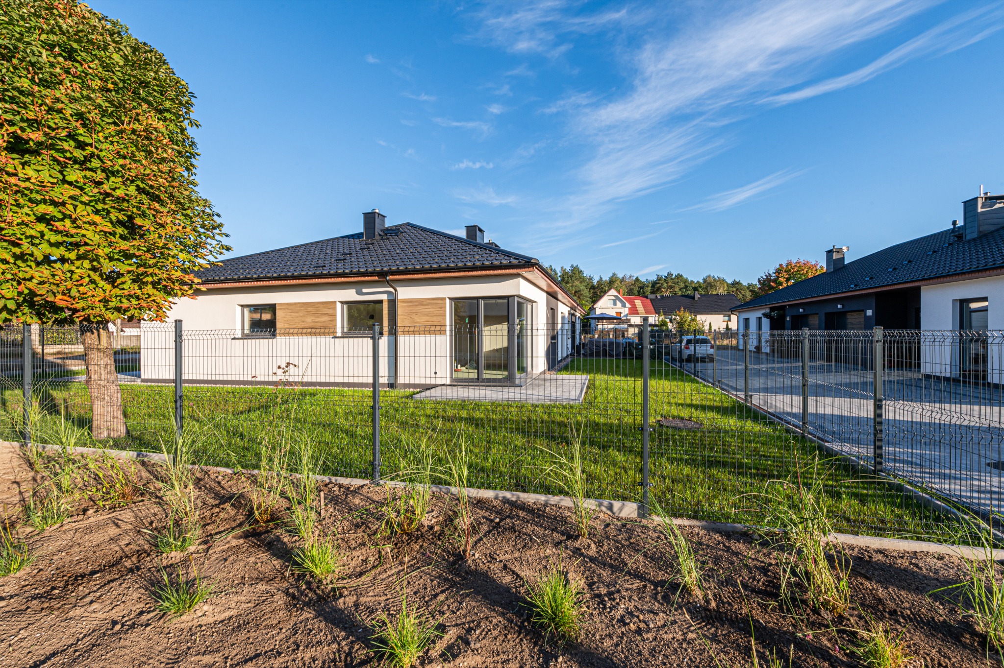 Nowoczesny dom jednorodzinny z szarą dachówką i jasną elewacją, otoczony zielonym trawnikiem i ogrodzeniem panelowym. W tle widoczne inne domy i błękitne niebo.