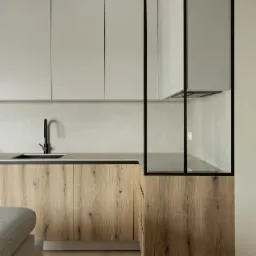 Nowoczesna kuchnia z drewnianymi szafkami dolnymi i gładkimi, szarymi szafkami górnymi, czarna armatura i szklana ścianka działowa z czarną ramą.