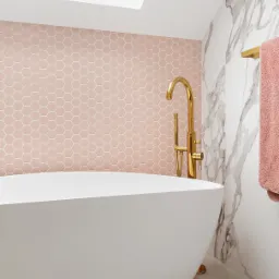 Nowoczesna łazienka z wolnostojącą białą wanną, złotą armaturą i różową mozaiką heksagonalną na ścianie akcentującej, doświetlona oknem dachowym.