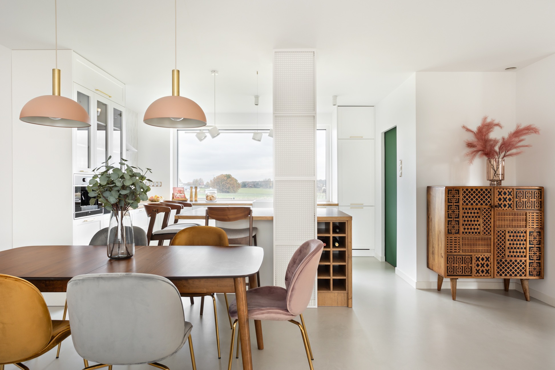 Jasna, przestronna jadalnia z drewnianym stołem otoczonym welurowymi krzesłami w odcieniach złota, szarości i różu; nad stołem wiszą lampy z różowymi kloszami; w tle widoczna otwarta kuchnia...
