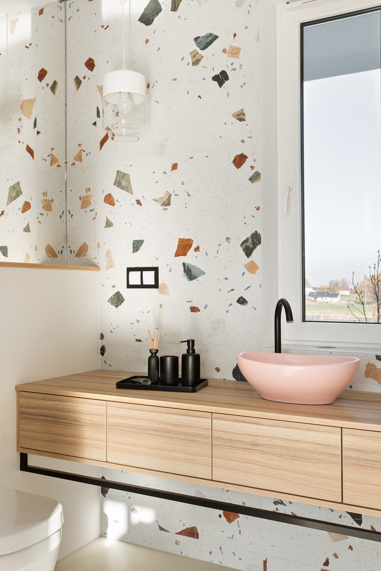 Nowoczesna łazienka z umywalką w kolorze pudrowego różu, armaturą w kolorze czarnym, blatem z jasnego drewna i ścianami wykończonymi płytkami imitującymi lastryko.