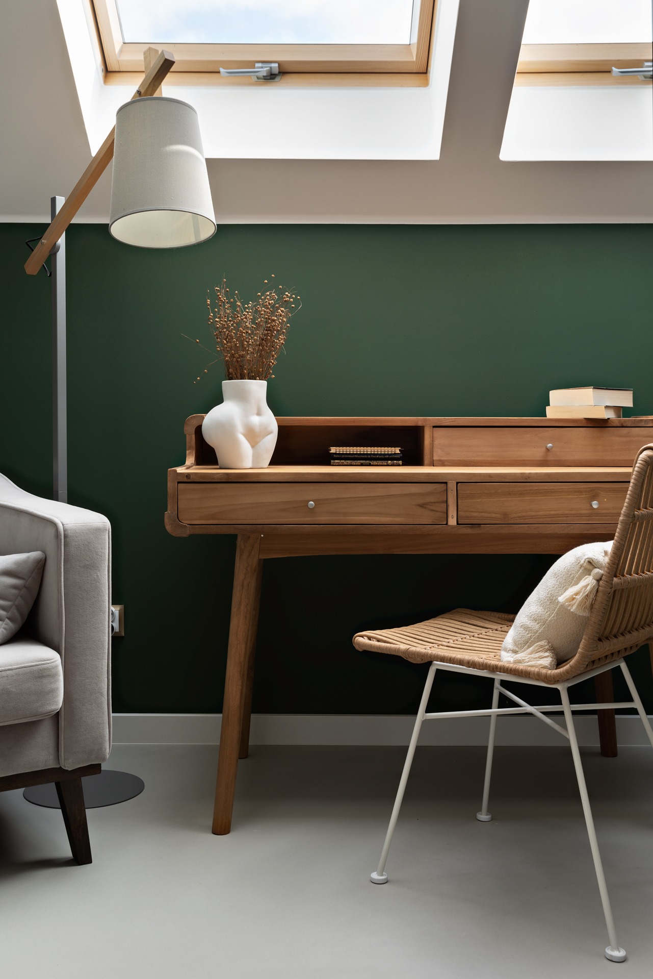 Drewniane biurko z krzesłem rattanowym i lampą podłogową w pokoju z zieloną ścianą i oknami dachowymi, dekorowane wazonem w kształcie kobiecego ciała i suchymi kwiatami.