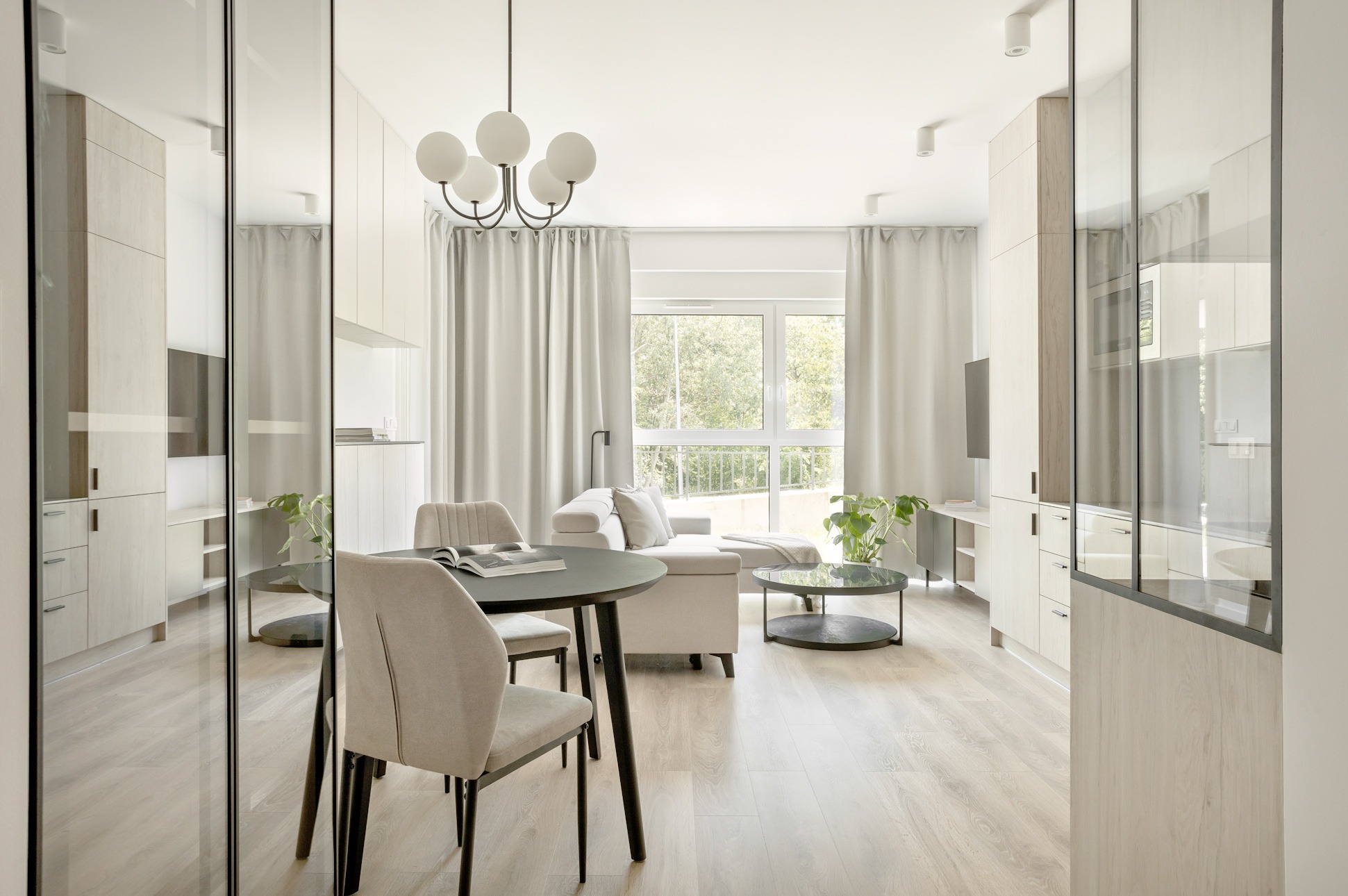 Jasne, przestronne wnętrze mieszkania z drewnianą podłogą, okrągłym stołem z krzesłami, sofą i przeszklonymi szafkami, oświetlone naturalnym światłem z okna z białymi zasłonami.