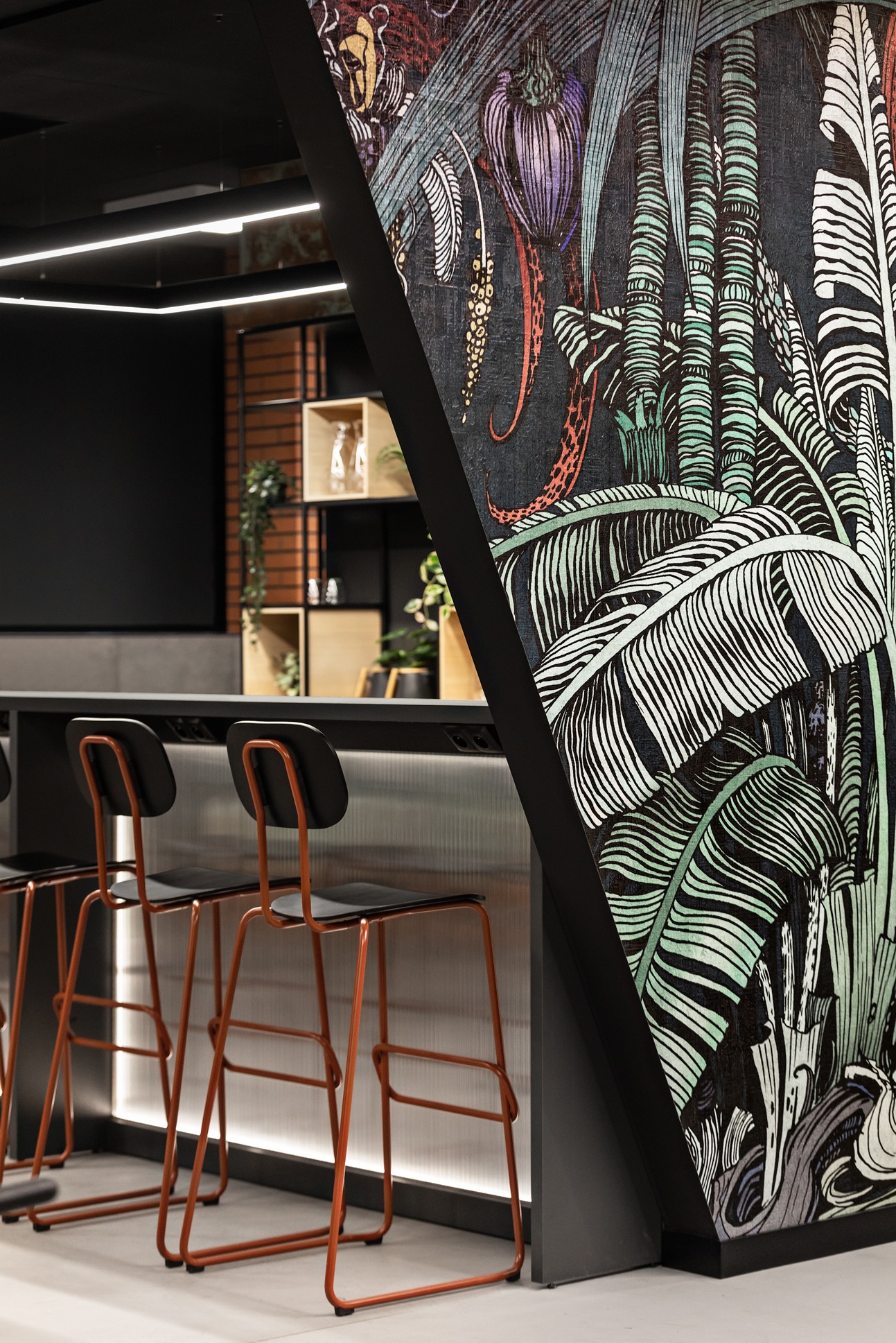 Nowoczesna przestrzeń biurowa z wysokimi krzesłami barowymi o pomarańczowych ramach, blatem z podświetlanego pleksi i ścianą ozdobioną ciemnym, tropikalnym wzorem roślinnym.