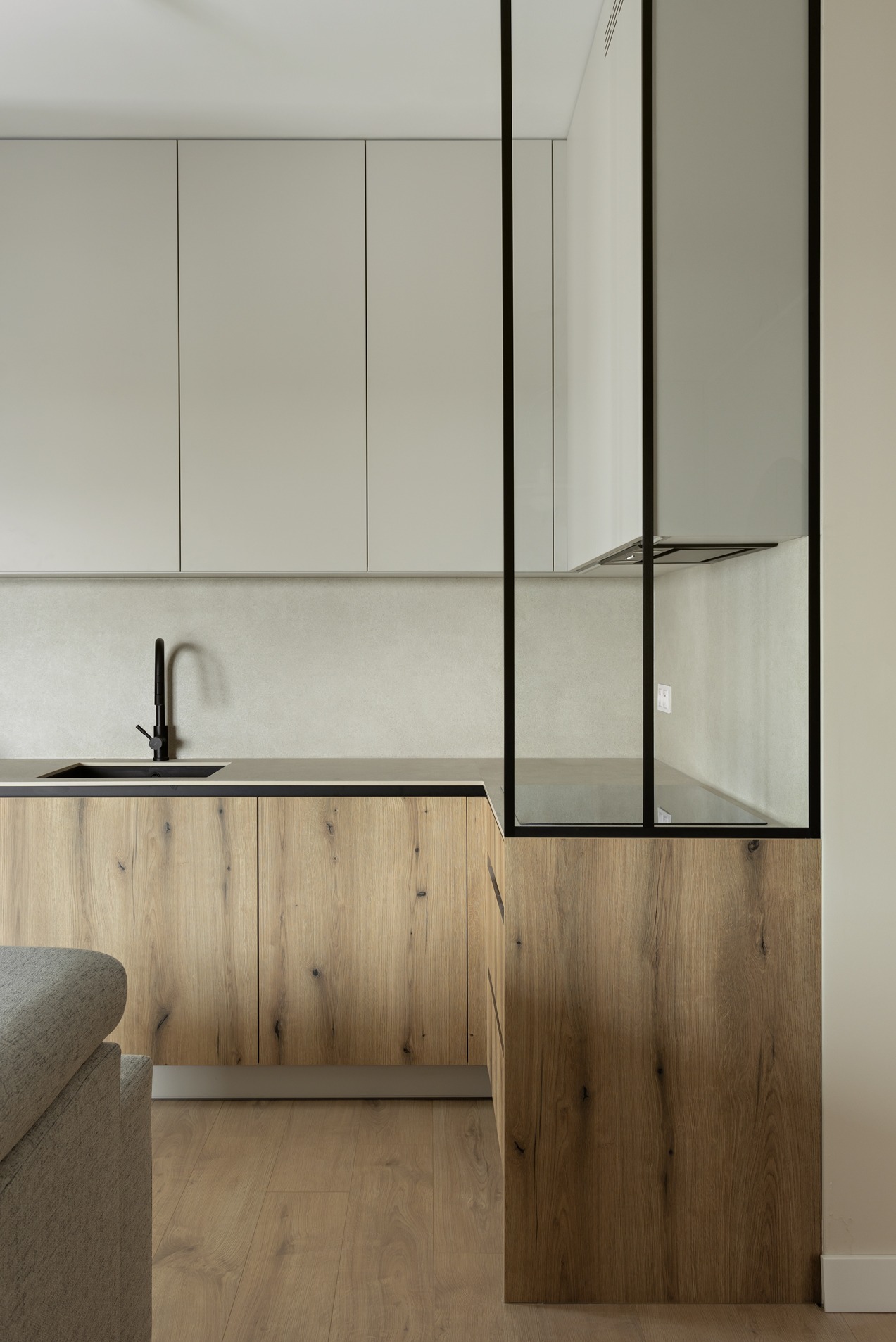 Nowoczesna kuchnia z drewnianymi szafkami dolnymi i gładkimi, szarymi szafkami górnymi, czarna armatura i szklana ścianka działowa z czarną ramą.