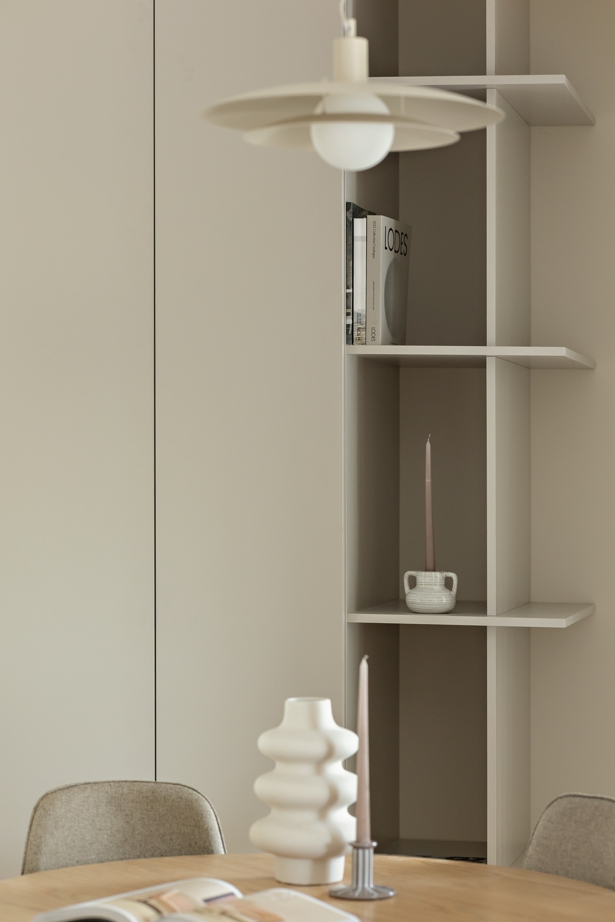 Minimalistyczne wnętrze: kremowa szafka z półkami, dekoracje (wazon, świece, książki LODES), lampa wisząca nad drewnianym stołem i krzesłem.