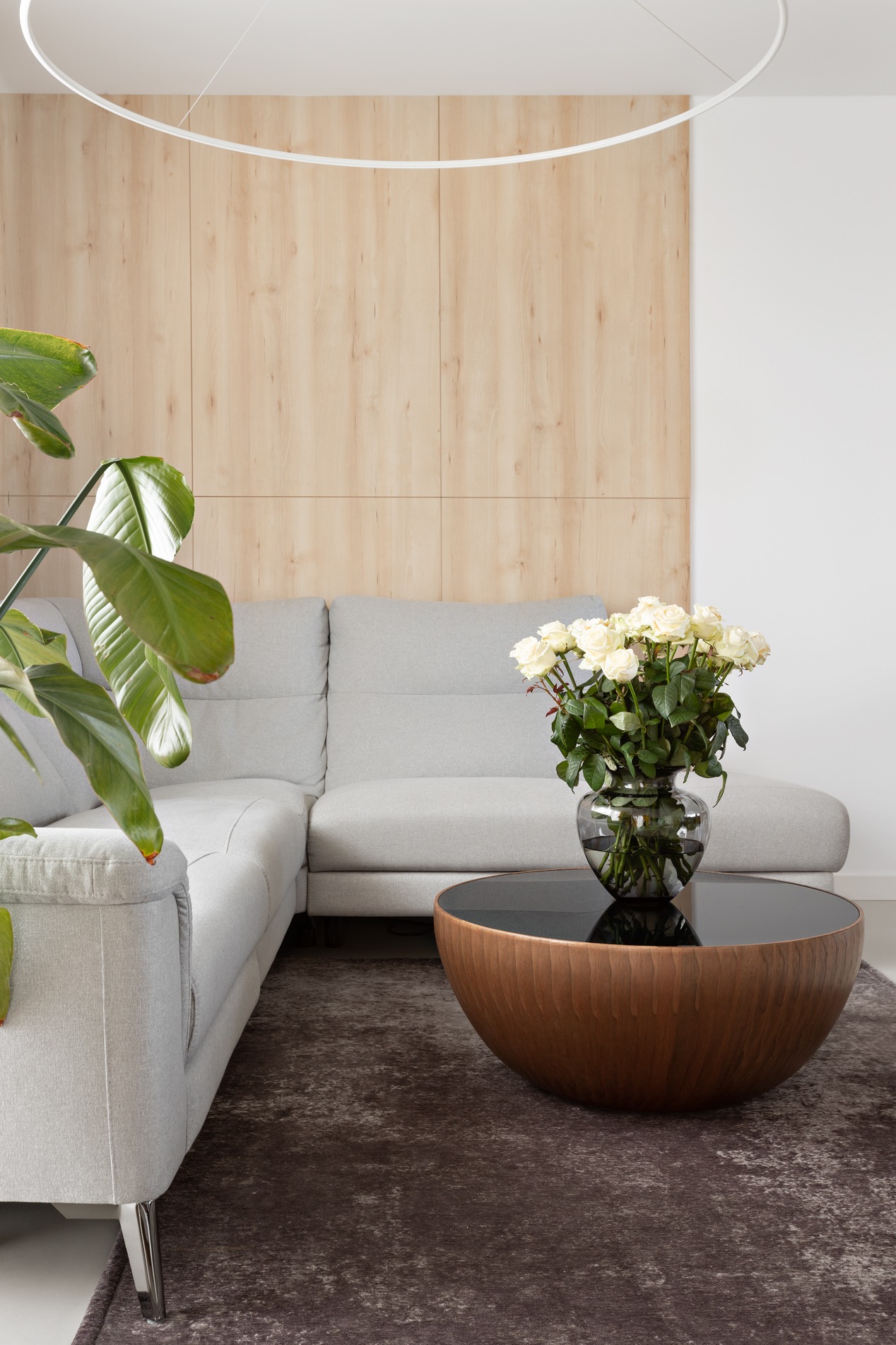 Jasnoszara sofa narożna z poduszkami, okrągły stolik kawowy z czarnym blatem i drewnianą podstawą, bukiet białych róż w szklanym wazonie, drewniana ściana w tle, minimalistyczna lampa sufitowa.