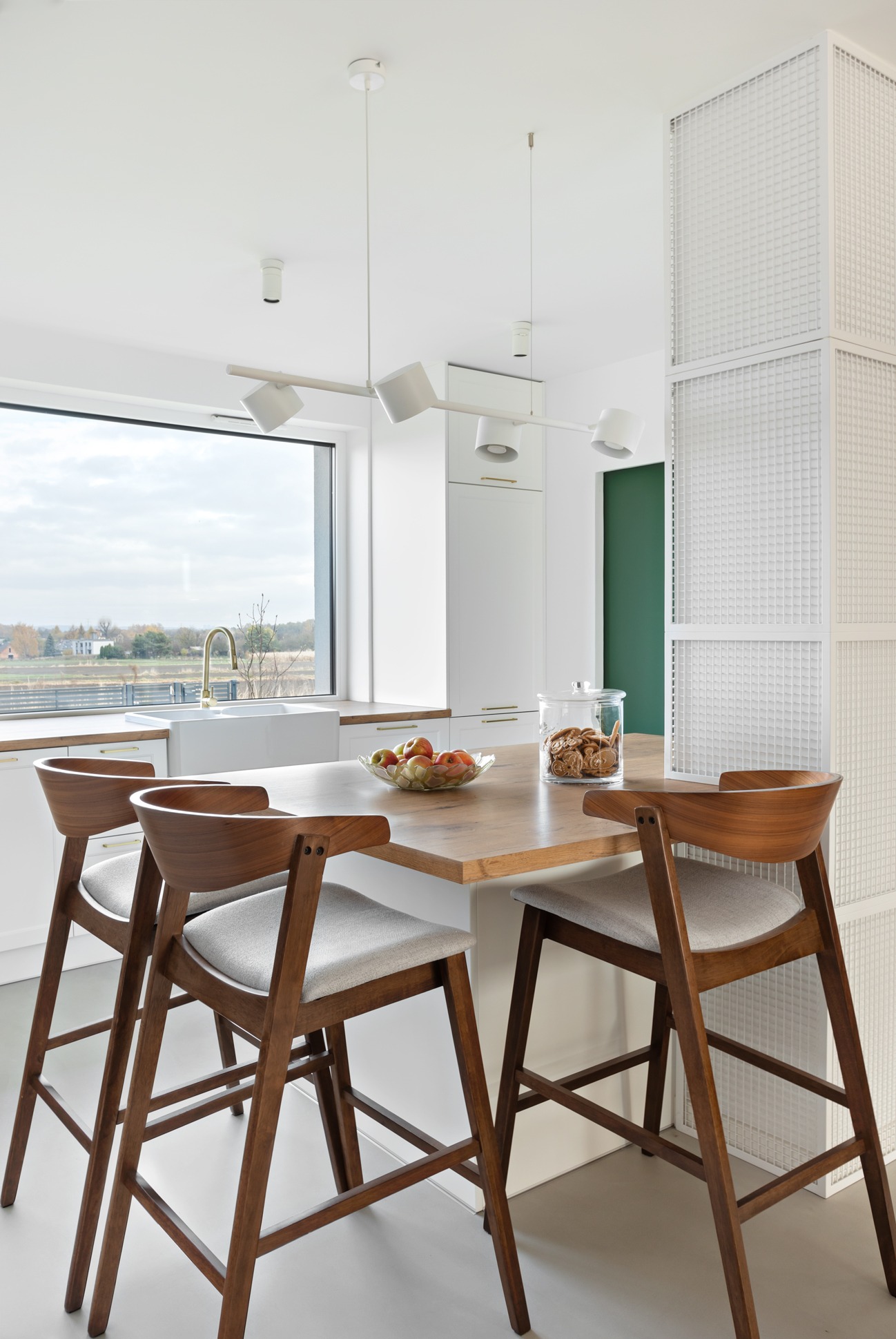 Jasna kuchnia z drewnianym blatem, wysokimi krzesłami barowymi i widokiem na krajobraz za oknem; na blacie szklana misa z jabłkami i słoik z ciasteczkami.