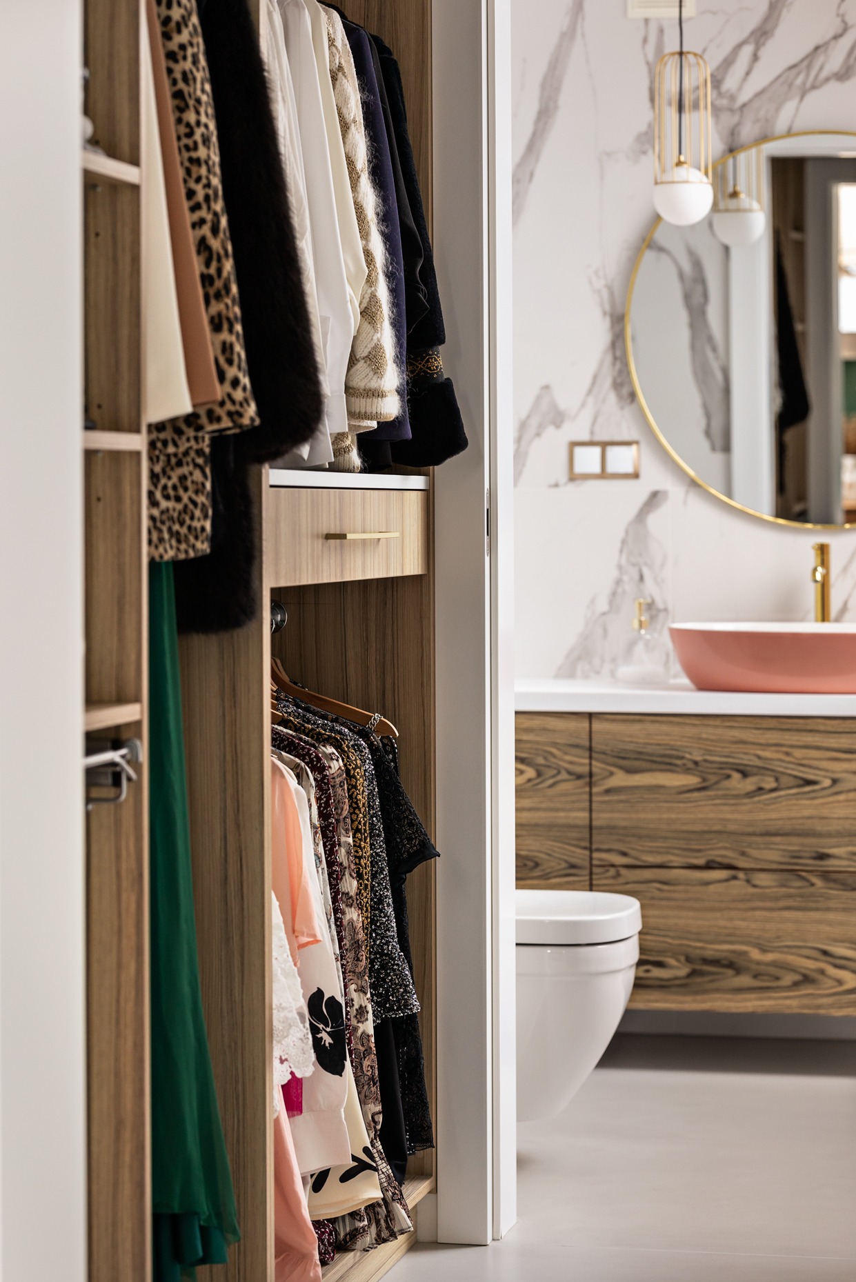 Widok z garderoby na łazienkę z marmurową ścianą, okrągłym lustrem w złotej ramie, umywalką w kolorze pudrowego różu i szafką z drewnianym fornirem.
