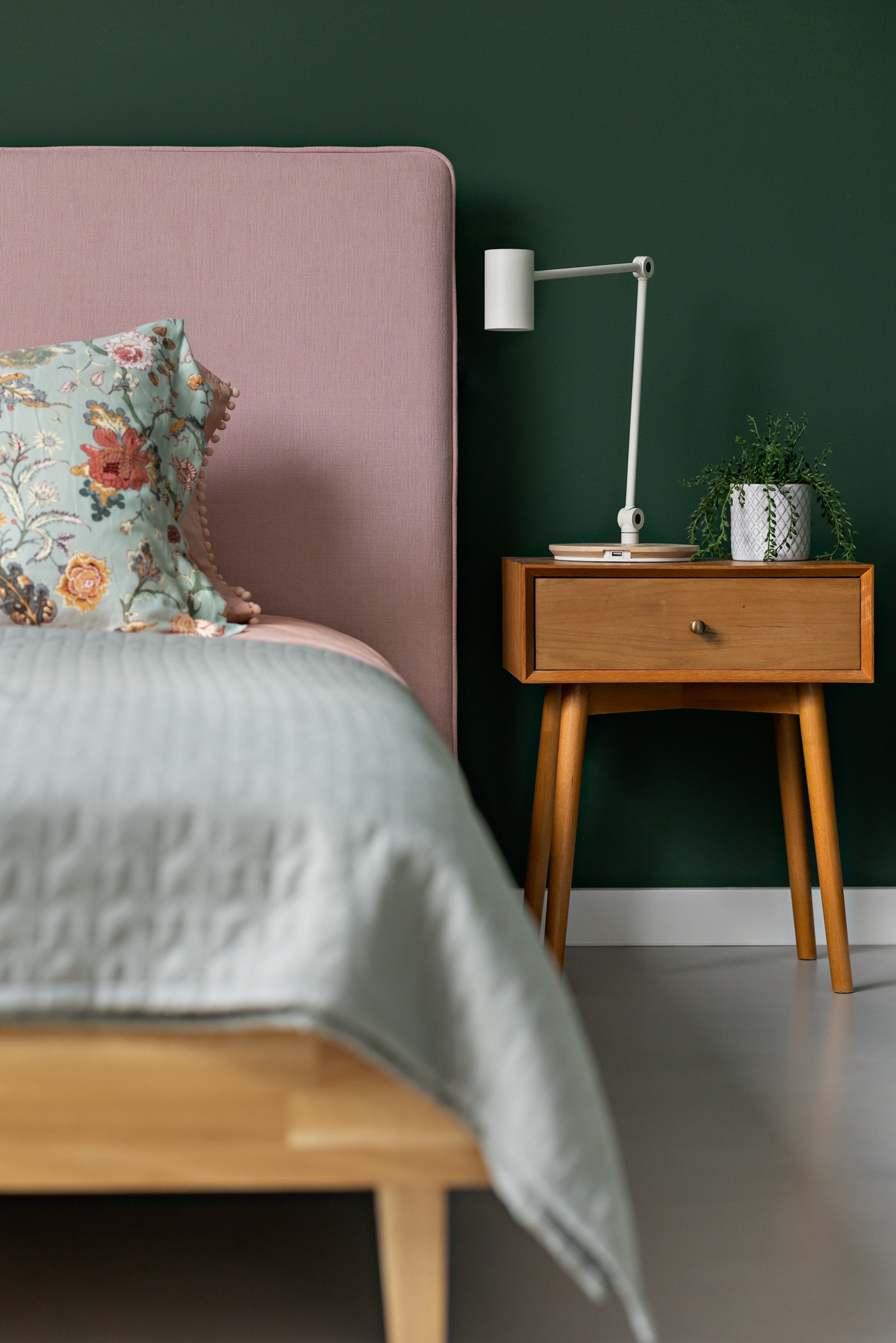 Fragment sypialni: łóżko z różowym zagłówkiem i kwiecistą poduszką, drewniana szafka nocna z lampką i doniczką na tle butelkowozielonej ściany.