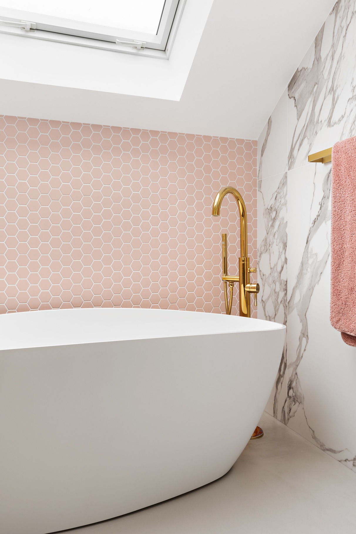 Nowoczesna łazienka z wolnostojącą białą wanną, złotą armaturą i różową mozaiką heksagonalną na ścianie akcentującej, doświetlona oknem dachowym.