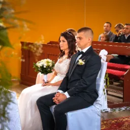 Para młoda siedzi w kościele podczas ceremonii ślubnej, panna młoda trzyma bukiet białych kwiatów, w tle goście weselni.