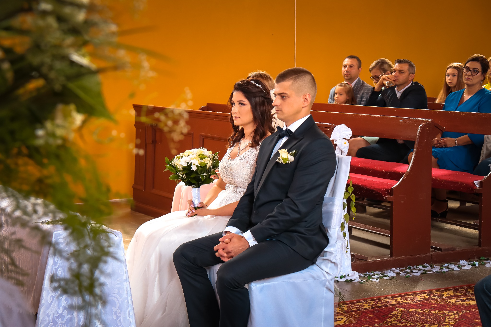 Para młoda siedzi w kościele podczas ceremonii ślubnej, panna młoda trzyma bukiet białych kwiatów, w tle goście weselni.