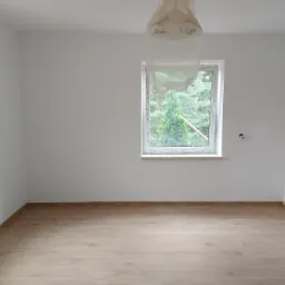 Pusty pokój po remoncie z drewnianą podłogą, świeżo pomalowanymi białymi ścianami, osłoniętą lampą i widokiem na zieleń za oknem.