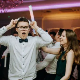 Rozbawiony mężczyzna w okularach i muszce, trzymający nad głową świecący pałeczkę i rekwizyt z papieru, otoczony przez uśmiechniętych gości na sali balowej z żyrandolem.