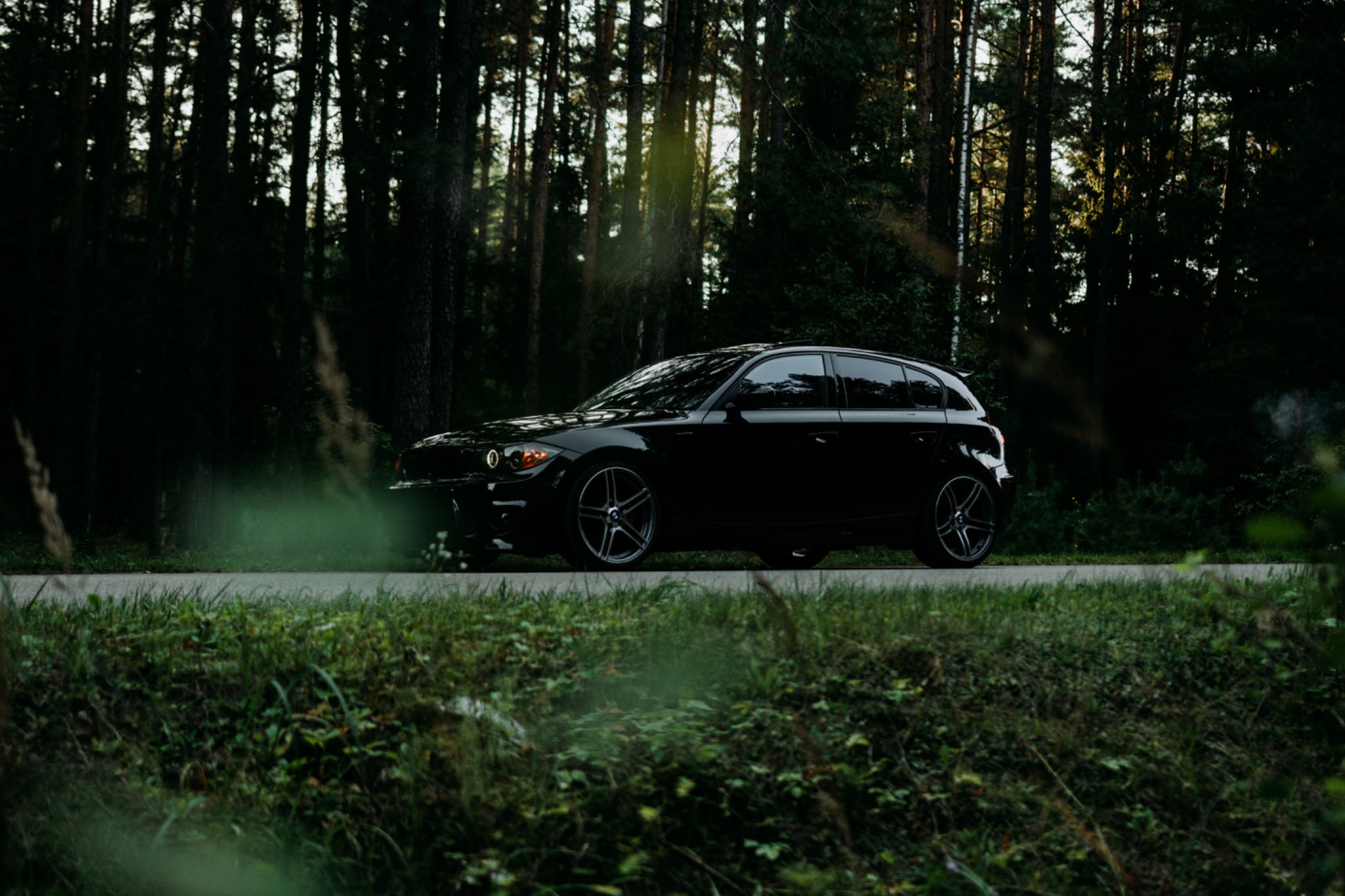 Czarny samochód typu hatchback zaparkowany na asfalcie z widocznym dymem z rury wydechowej na tle ciemnego lasu iglastego, z zieloną roślinnością w pierwszym planie.