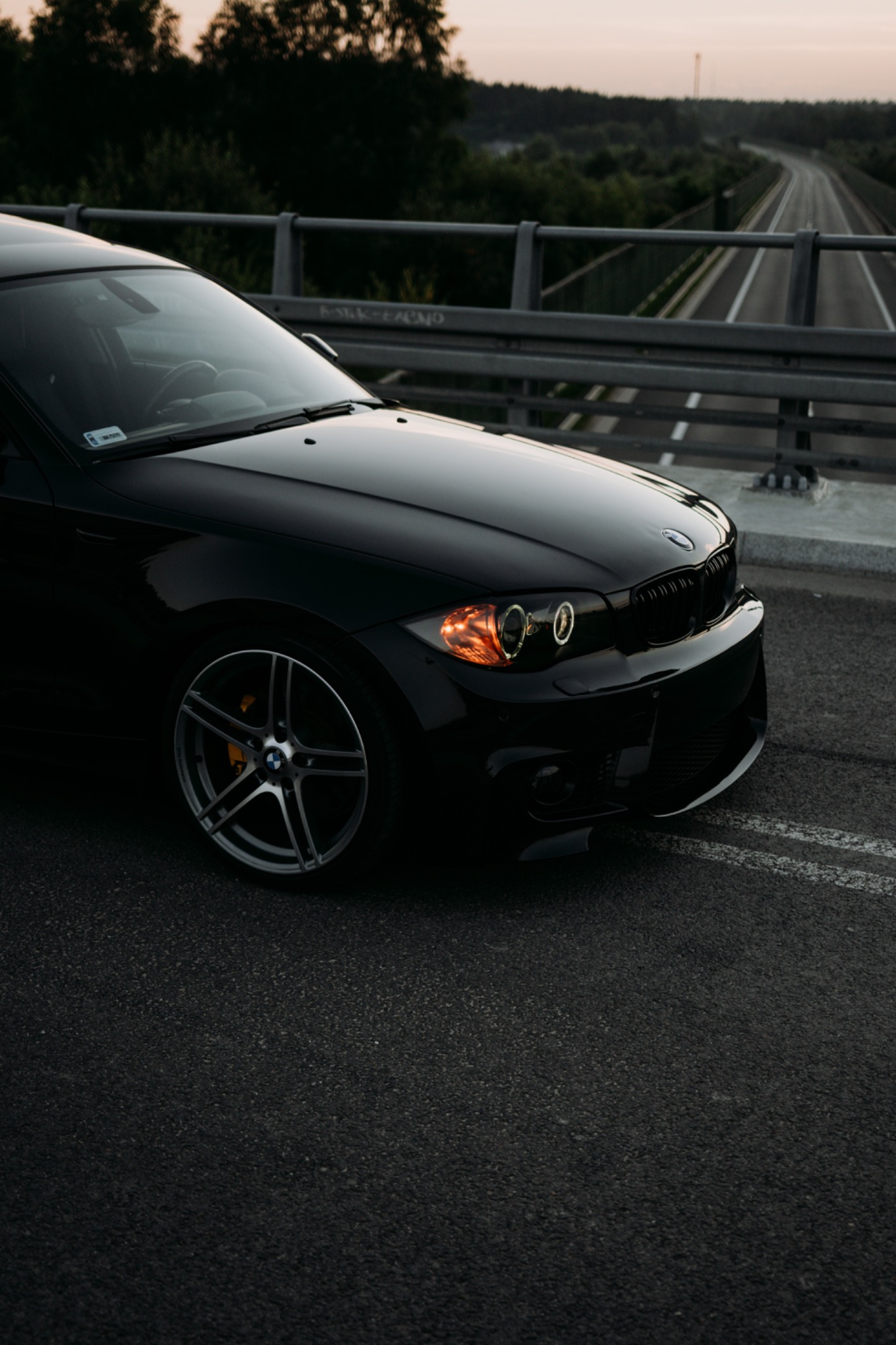 Częściowy widok czarnego samochodu na moście o zmierzchu, z widoczną drogą w tle i drzewami w oddali.