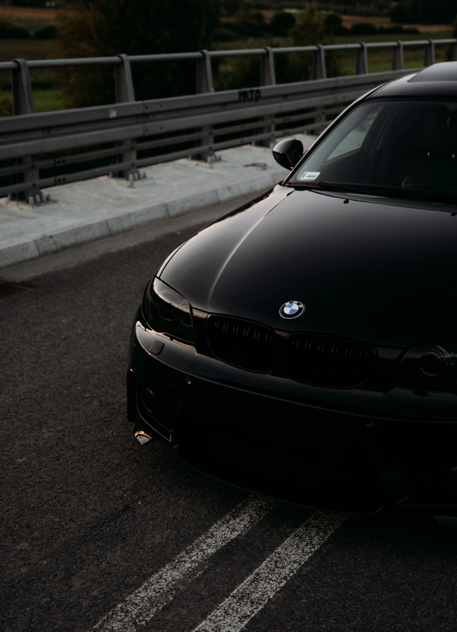 Częściowy widok z przodu czarnego samochodu BMW na tle betonowego mostu z metalową barierką w słoneczny dzień.