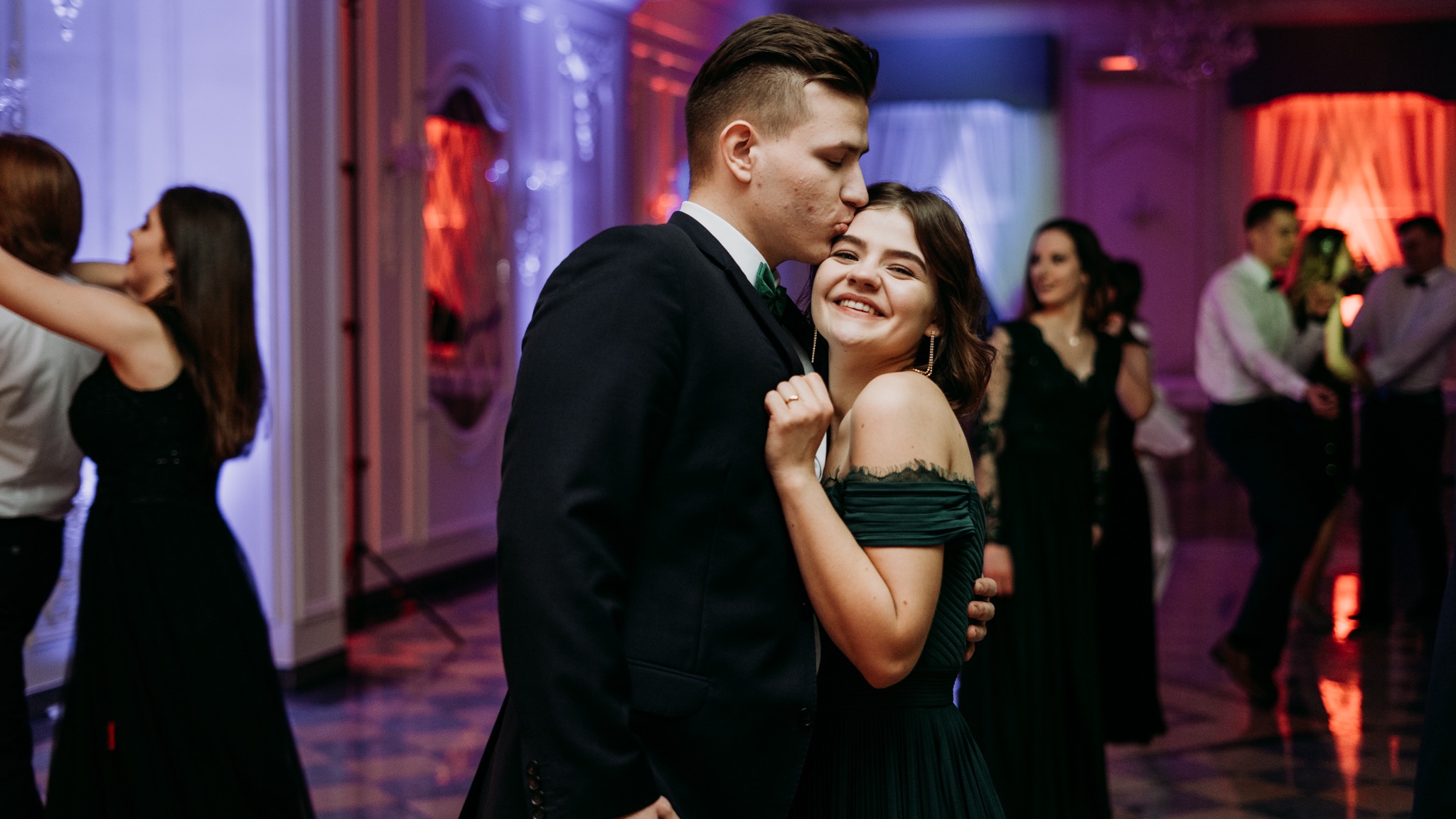 Para w eleganckich strojach tańczy na balu w Białymstoku, mężczyzna całuje kobietę w czoło, w tle inni goście bawią się na parkiecie.