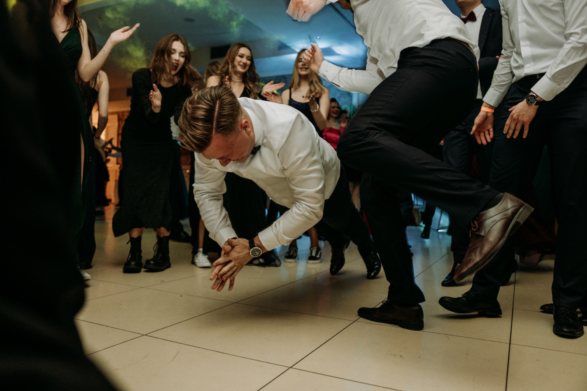 Mężczyzna w białej koszuli i czarnych spodniach tańczy nisko przy ziemi na parkiecie podczas imprezy, otoczony przez rozbawionych gości.