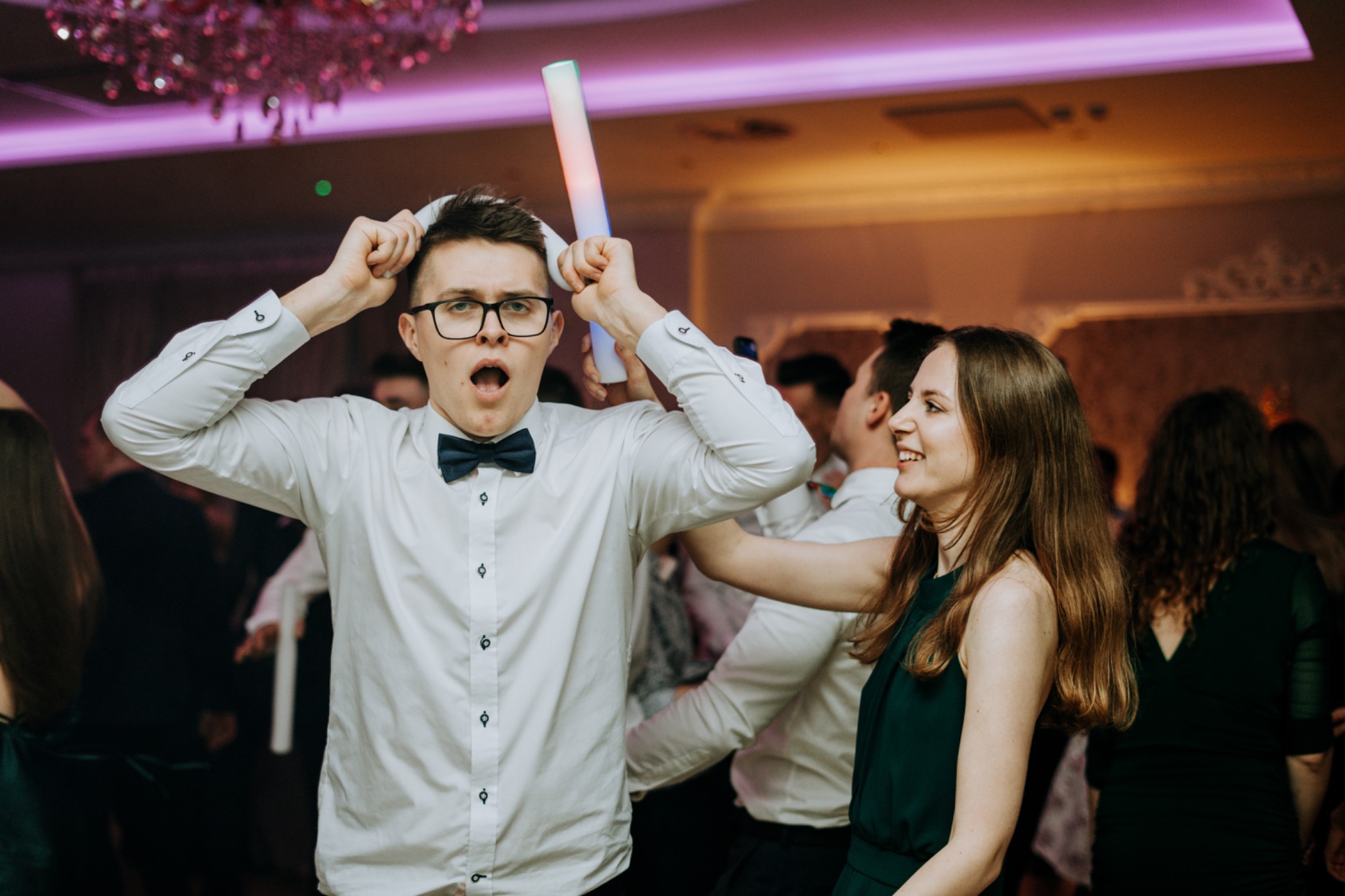 Rozbawiony mężczyzna w okularach i muszce, trzymający nad głową świecący pałeczkę i rekwizyt z papieru, otoczony przez uśmiechniętych gości na sali balowej z żyrandolem.