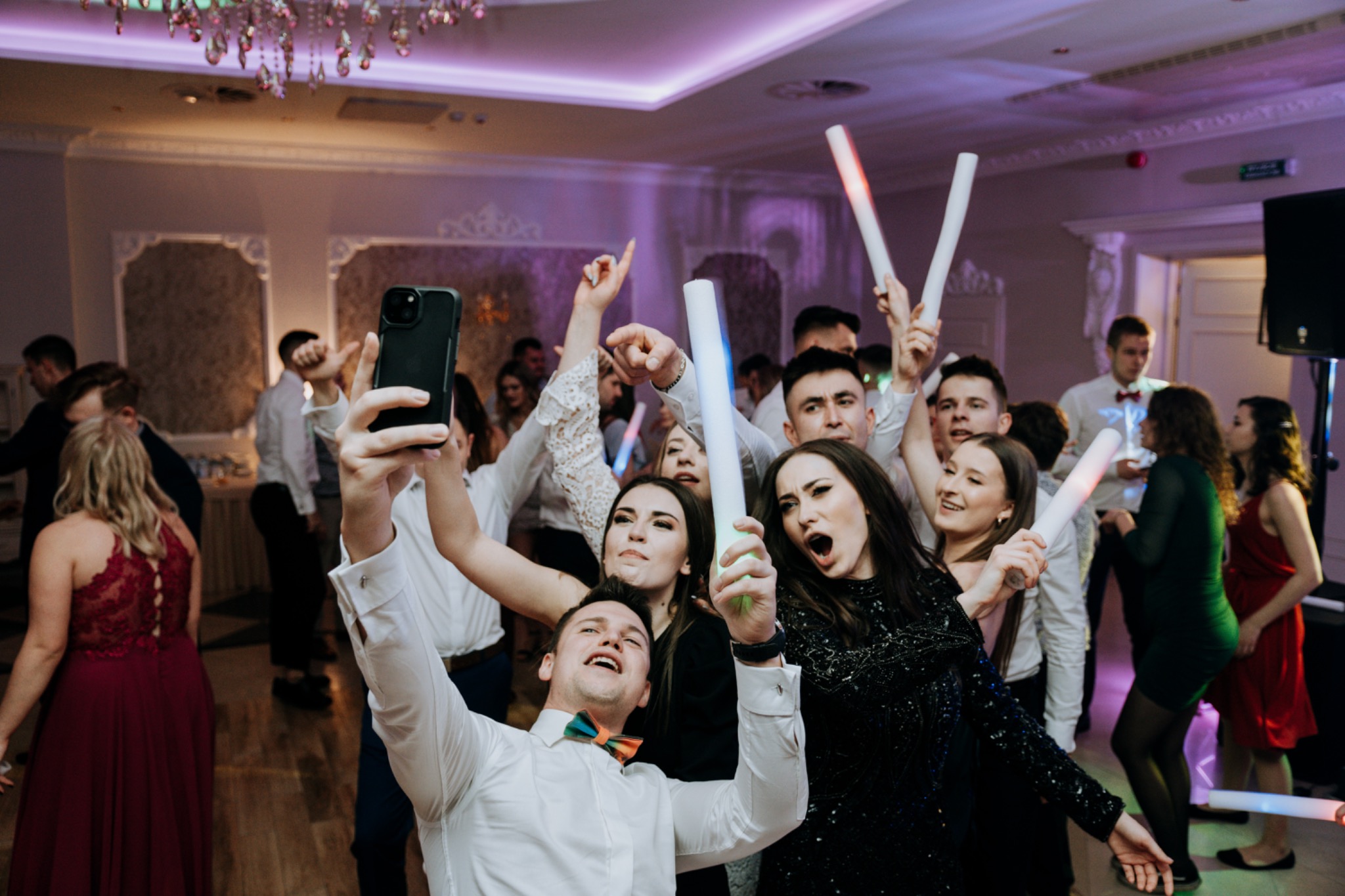 Grupa rozbawionych gości weselnych robiąca selfie telefonem, trzymająca świetlne pałeczki, w tle dekoracje sali i kryształowy żyrandol.