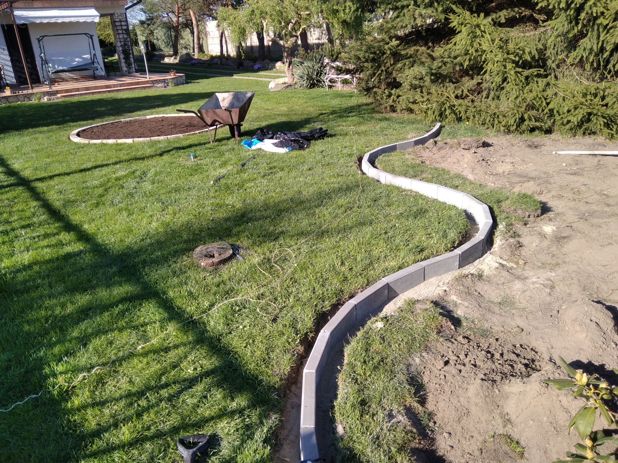 Krawężniki z szarej kostki betonowej wyznaczają ścieżkę w trawniku, widoczna taczka, ziemia przygotowana pod rabatę, rozłożone narzędzia i materiały budowlane na trawie.