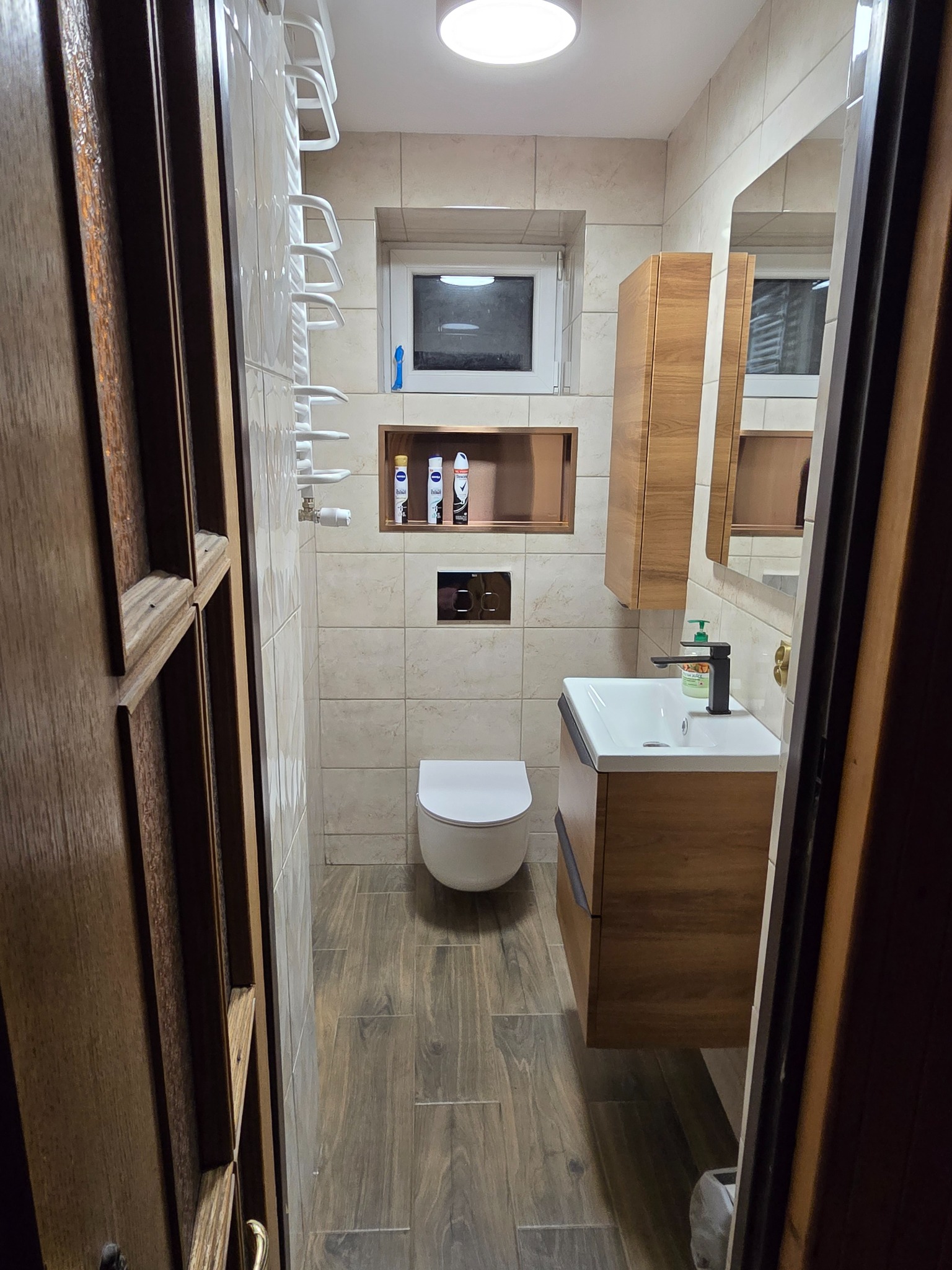 Nowoczesna łazienka z beżowymi płytkami, drewnopodobną podłogą, wiszącą szafką z umywalką i wnęką na kosmetyki nad toaletą, widziana zza uchylonych, drewnianych drzwi.