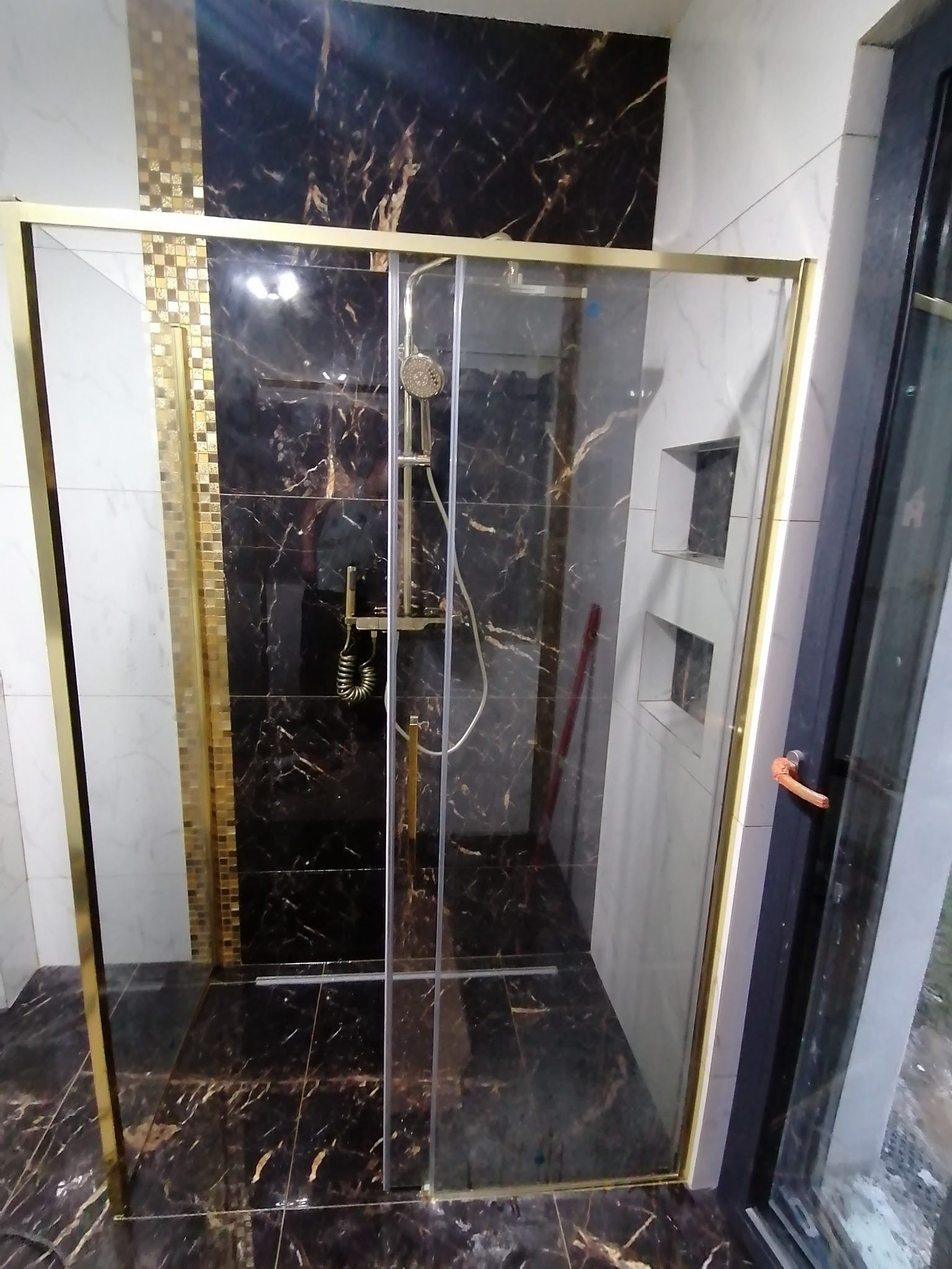 Kabina prysznicowa ze złotymi ramami na tle płytek imitujących czarny marmur i białych płytek z wnękami, widok z zewnątrz.